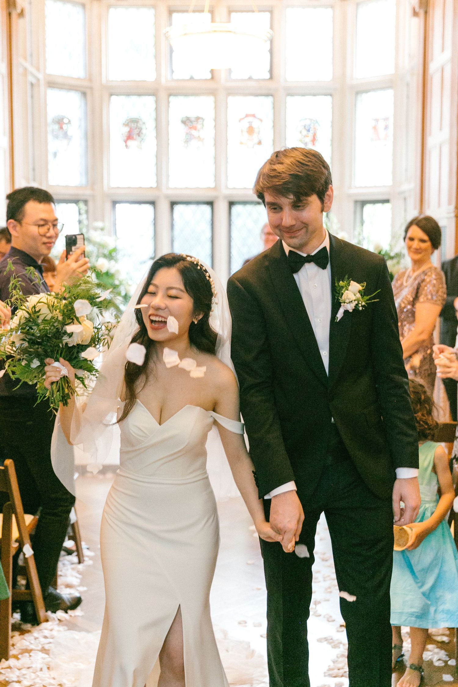 Joyful newlyweds walk back down the aisle as guests toss white flower petals in elegant venue.
