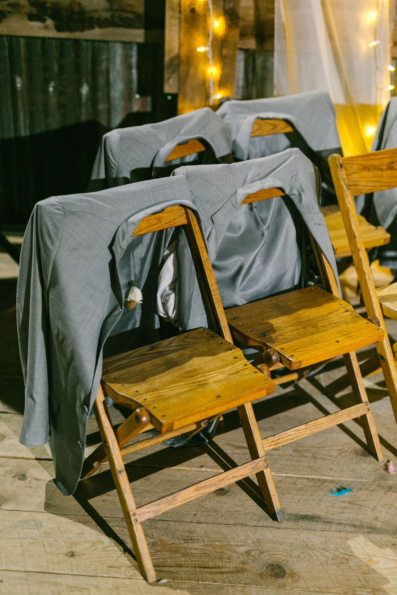 Wooden folding chairs with gray covers arranged at a rustic wedding venue with string lights in the background.