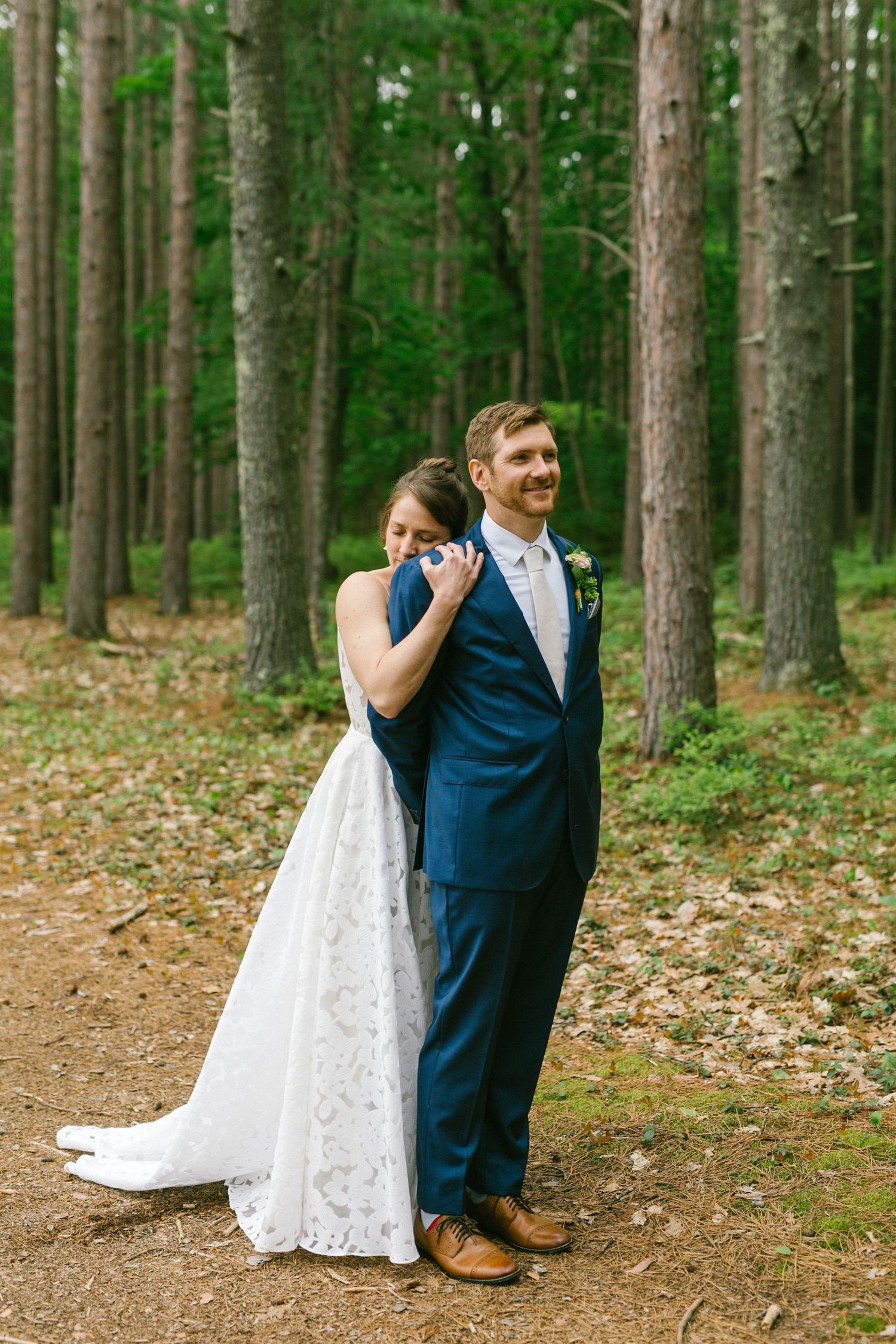 A wedding couple standing together in a serene forest setting with tall pine trees and a dirt path.