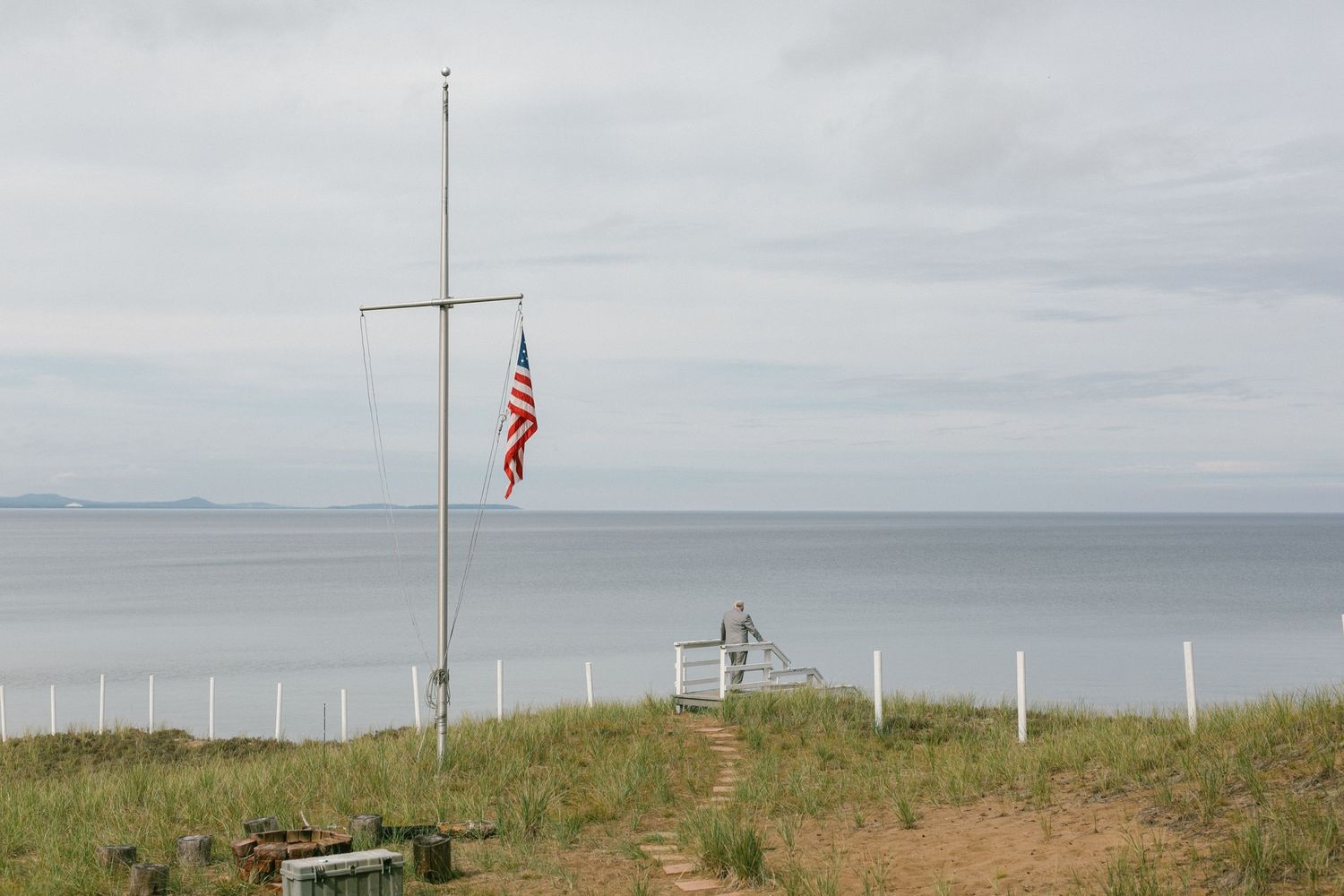 An American flag flies on a coastal hilltop overlooking a misty gray ocean and grassy dunes.