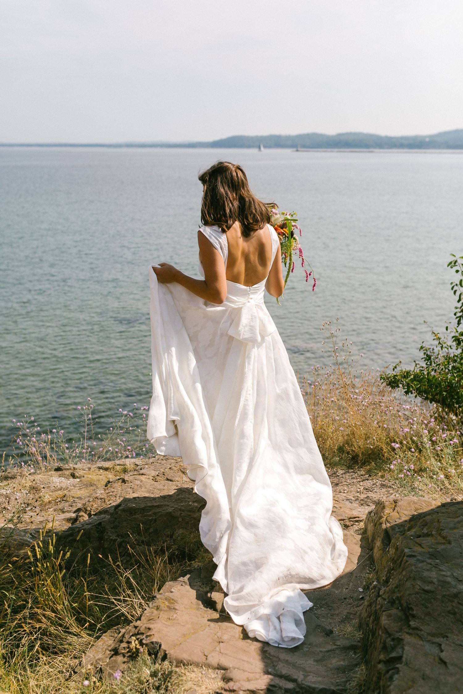 Bride in flowing white dress walks along coastal cliff edge with ocean view.