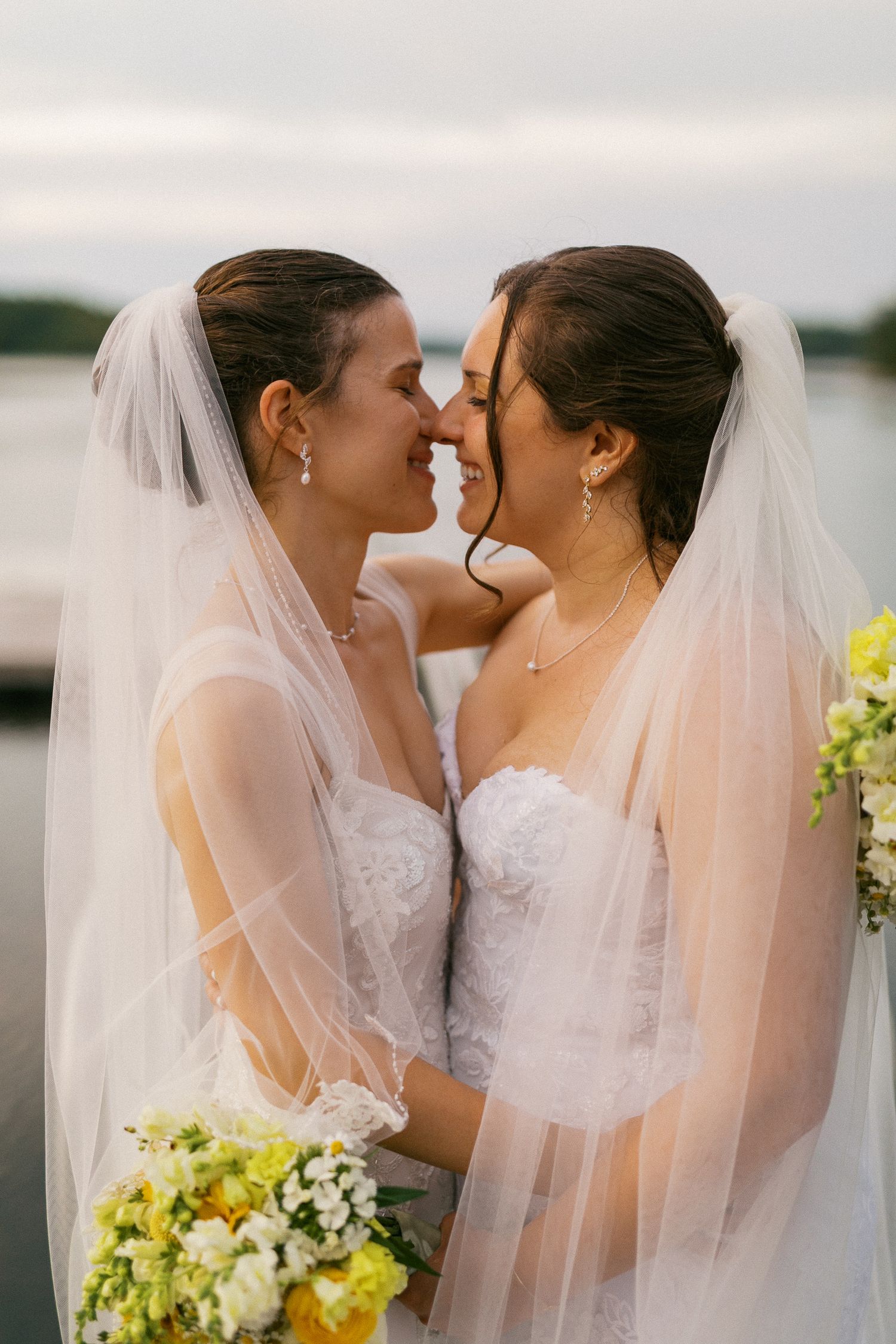 Two brides in white strapless gowns and veils touch foreheads, smiling, holding yellow bouquets near a lakeside.