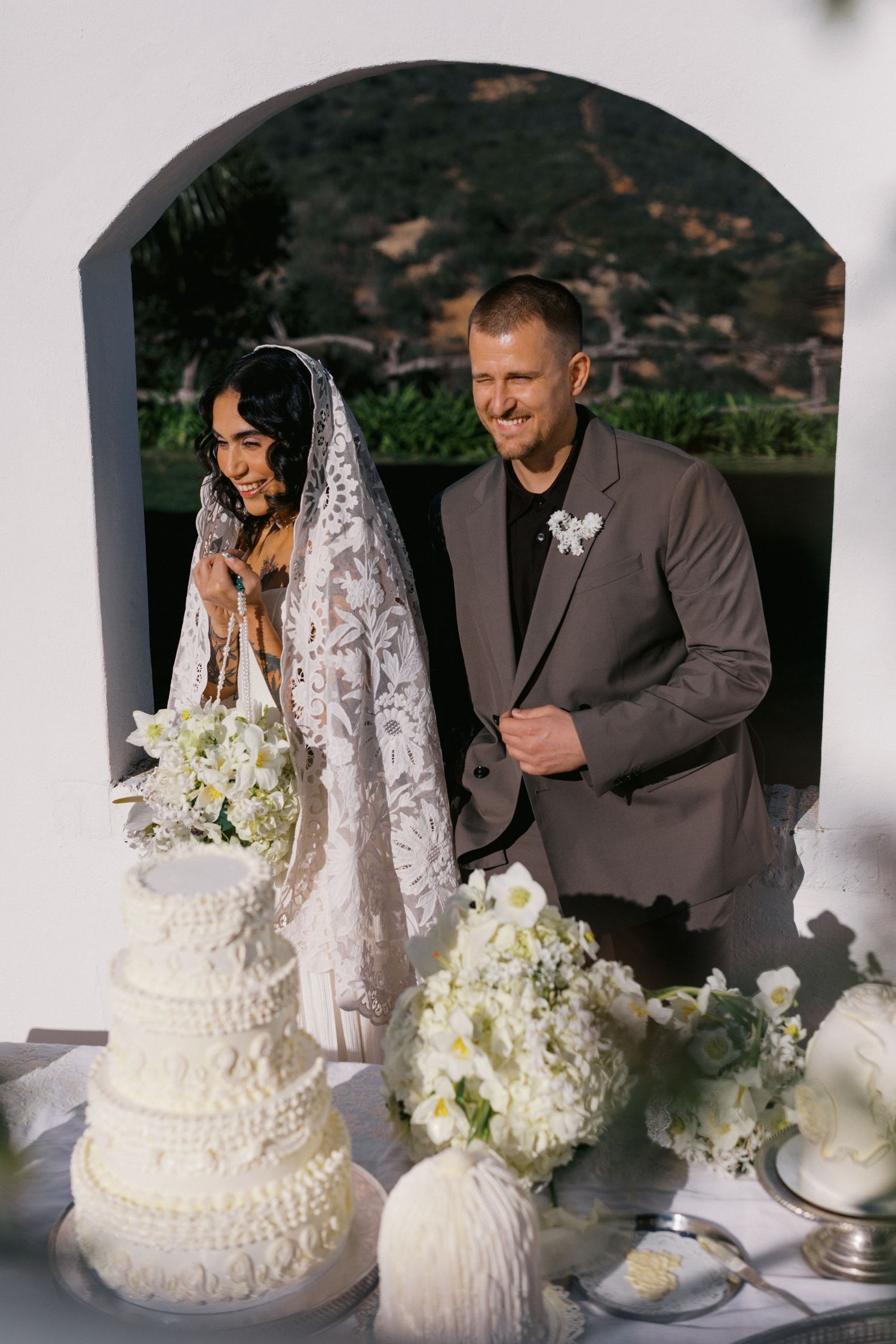 Bride and groom smiling beside a white tiered wedding cake with floral arrangements outdoors.