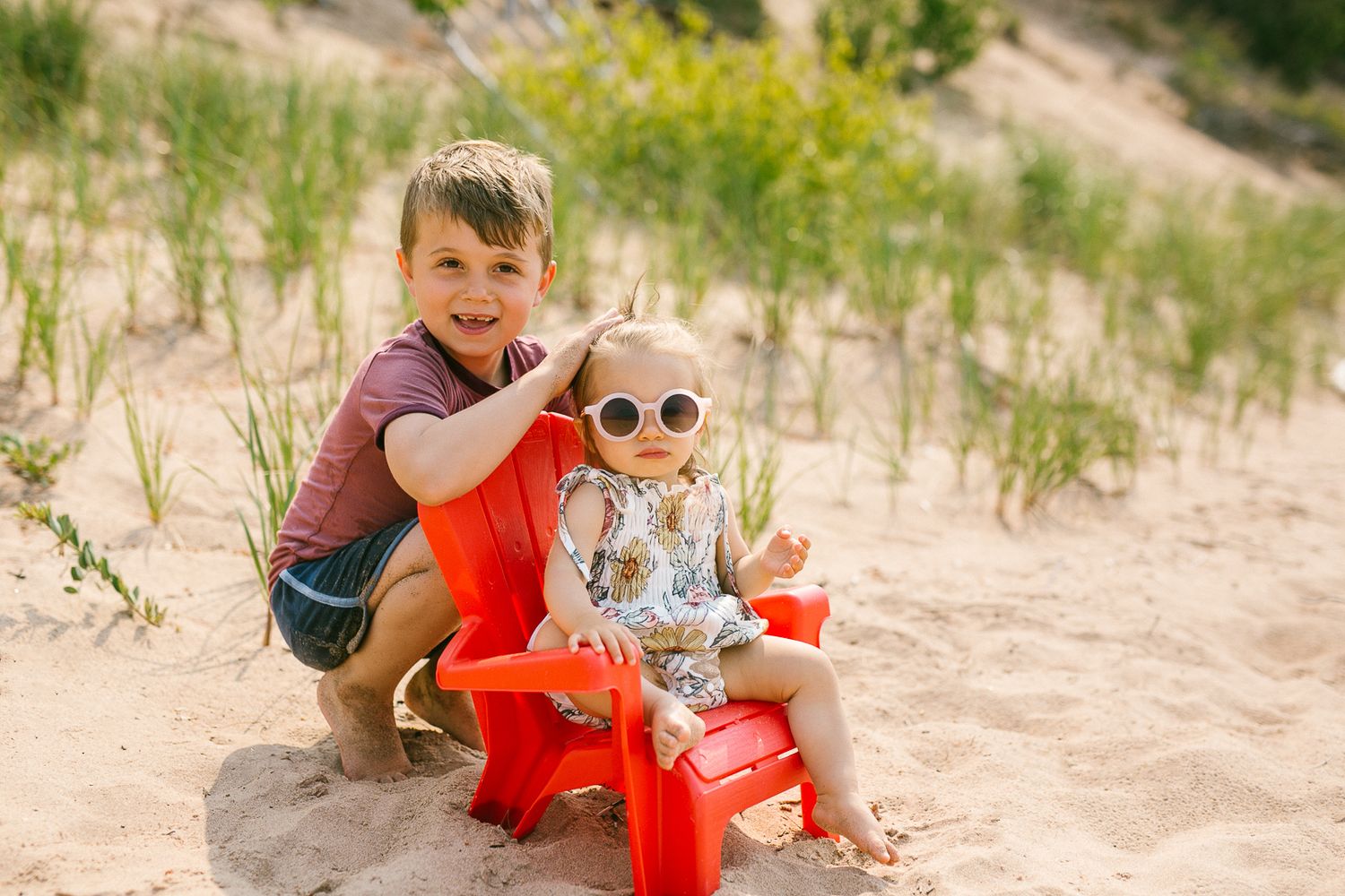 Happy boy crouches beside a smiling toddler in sunglasses sitting on a red chair in sandy dunes.