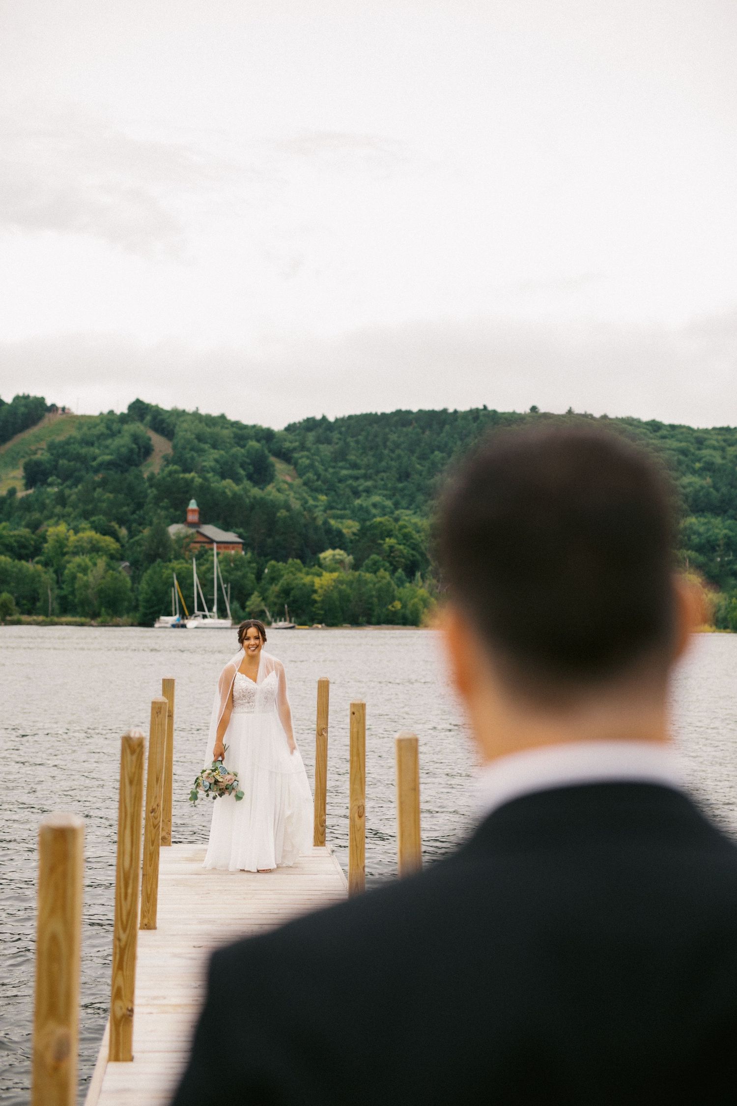 Bride walking on lakeside dock toward groom during romantic first look moment.