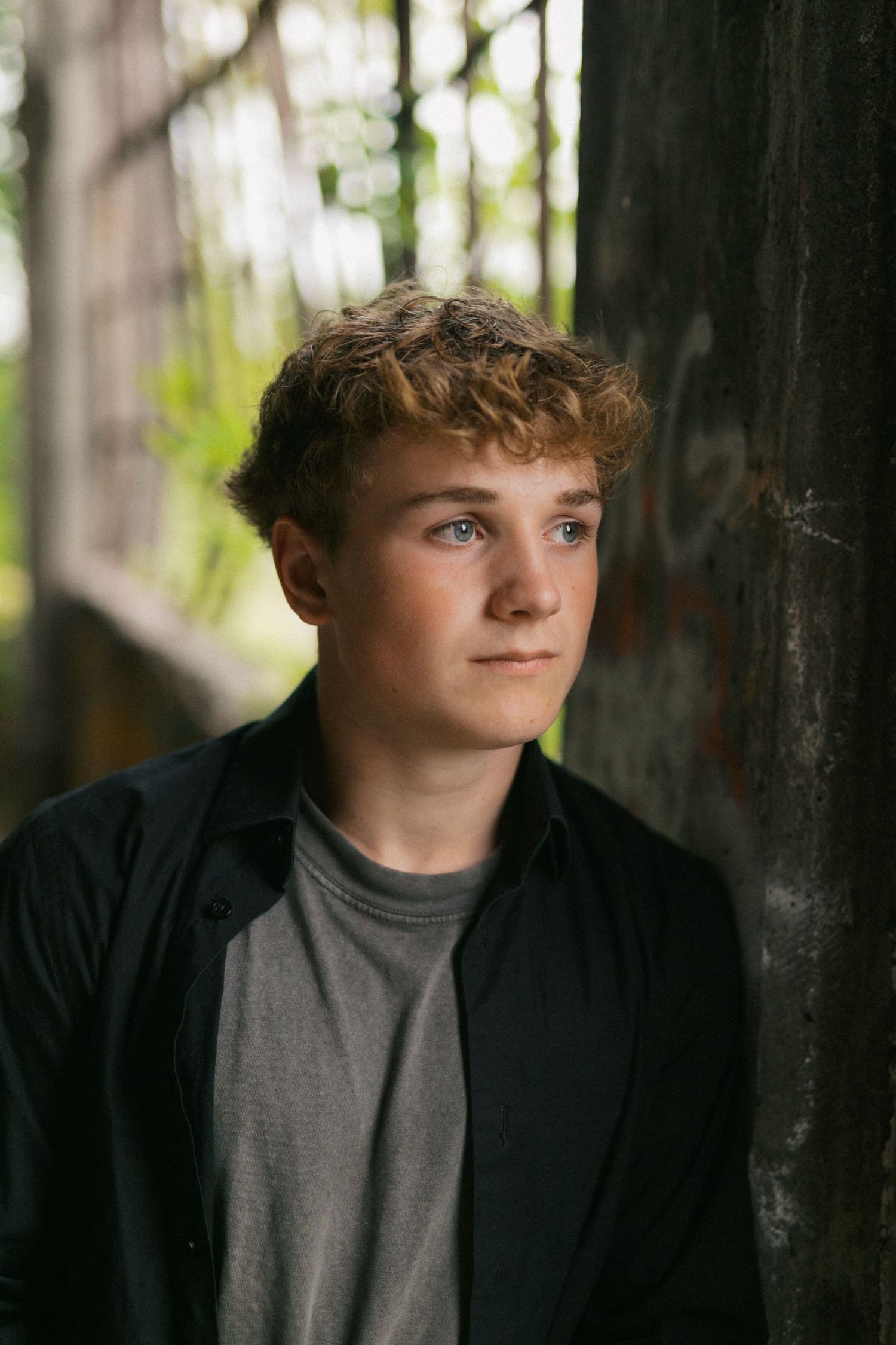 Teen boy with curly hair in black shirt leaning against concrete wall outdoors.