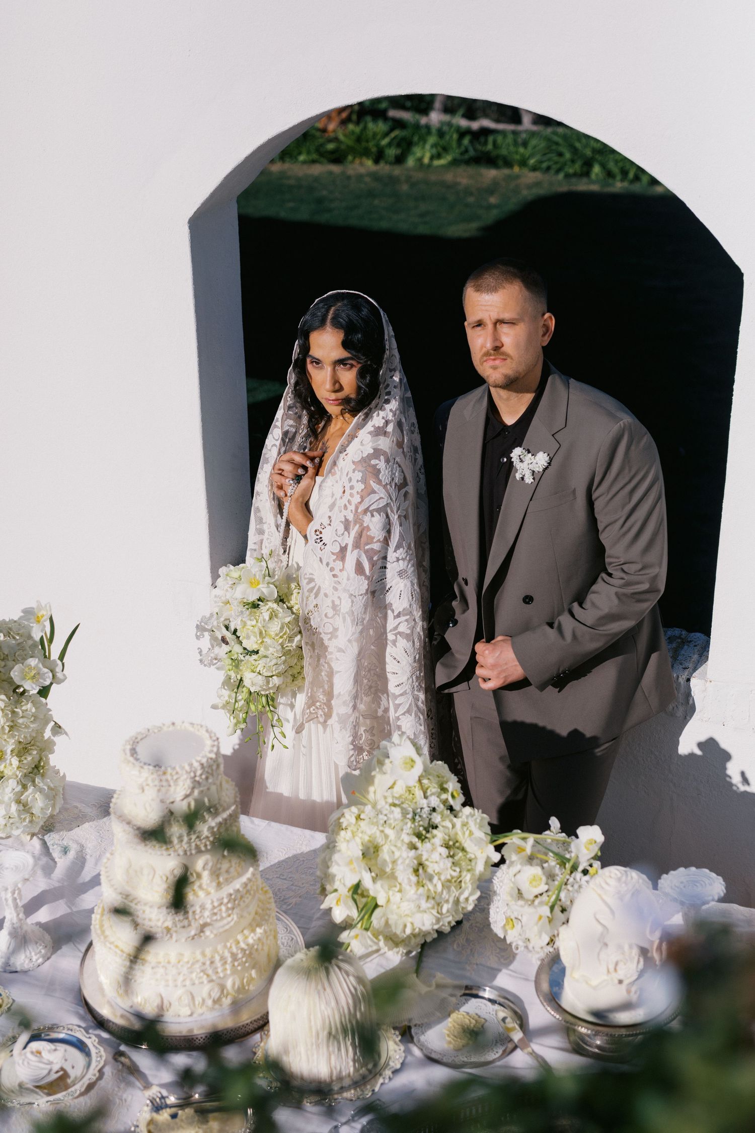 Bride and groom stand in arched doorway behind white floral wedding cake display at outdoor ceremony.