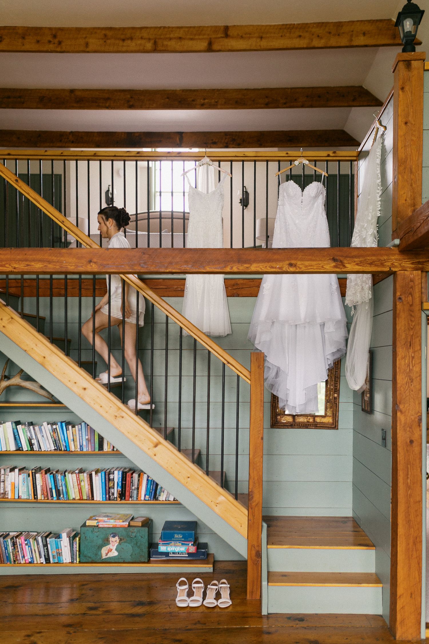Wedding dresses hanging on staircase railing in rustic barn venue with built-in bookshelf and bridal shoes on steps.