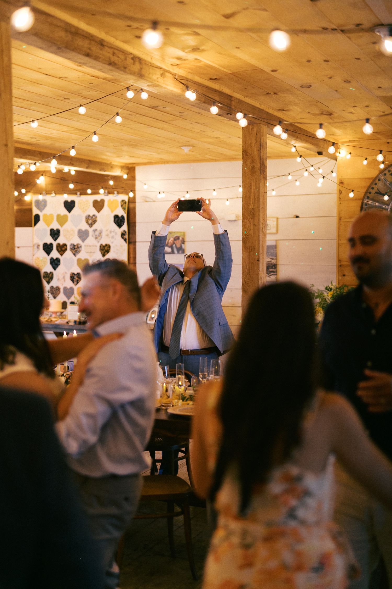 Guests celebrate at a rustic wedding reception with string lights and a heart backdrop while a man photographs the event.
