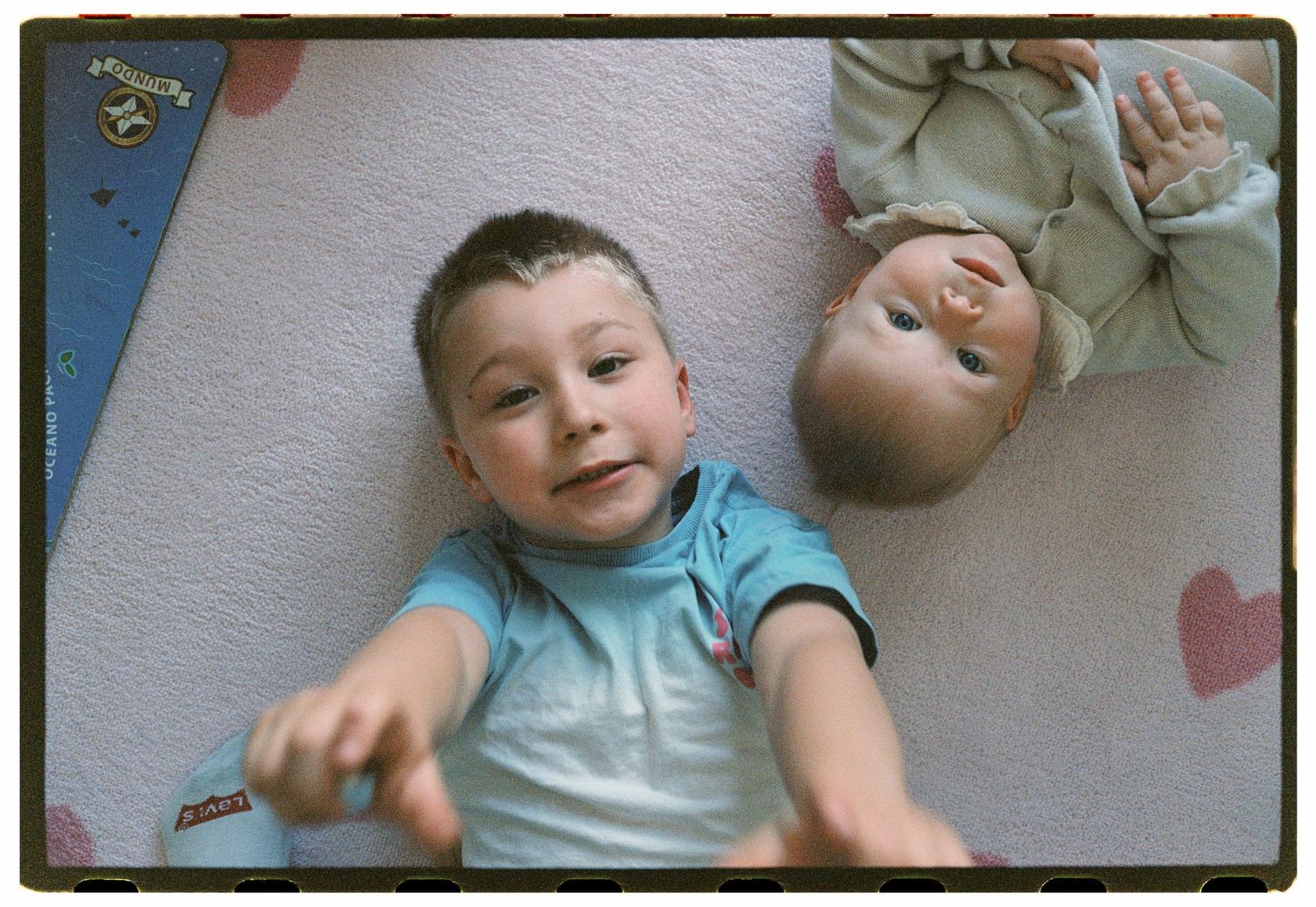 Young boy lying on a pink heart-patterned floor next to a baby doll, reaching toward the camera.