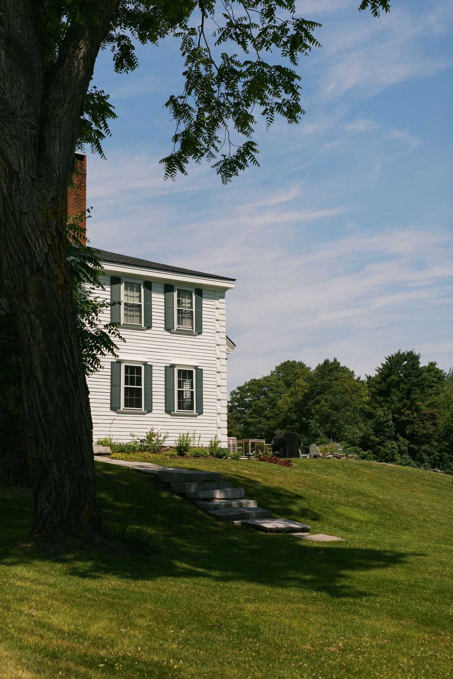 White colonial farmhouse with green shutters on a sunny hill, framed by a large tree and lush green lawn.