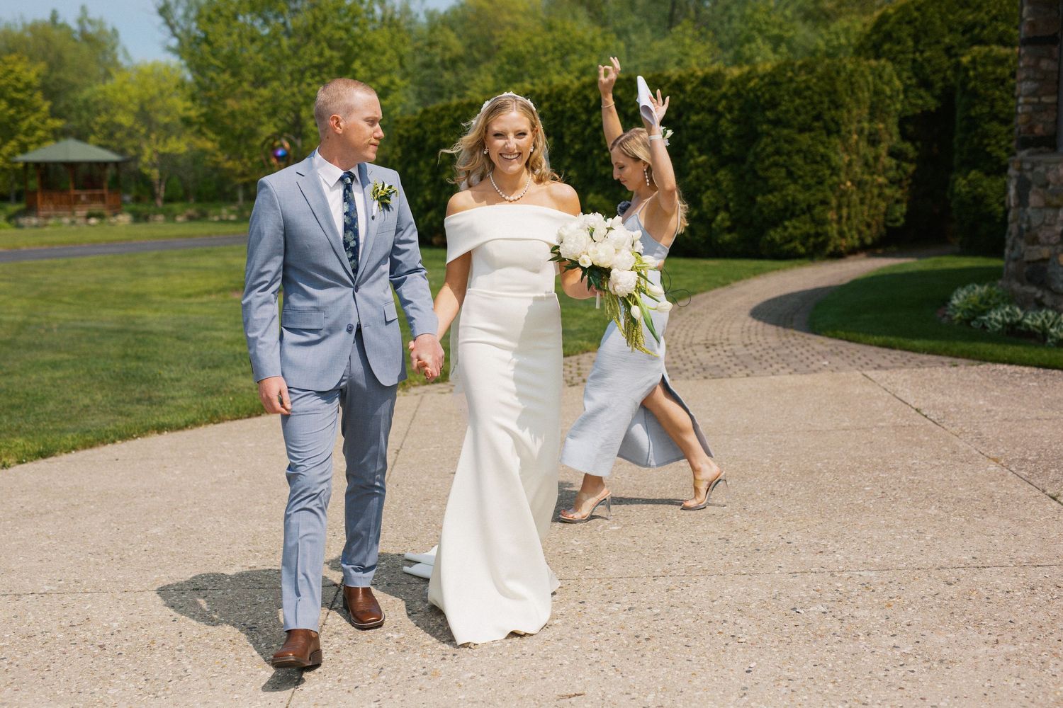 Wedding party walking together on gravel path surrounded by green landscaping.
