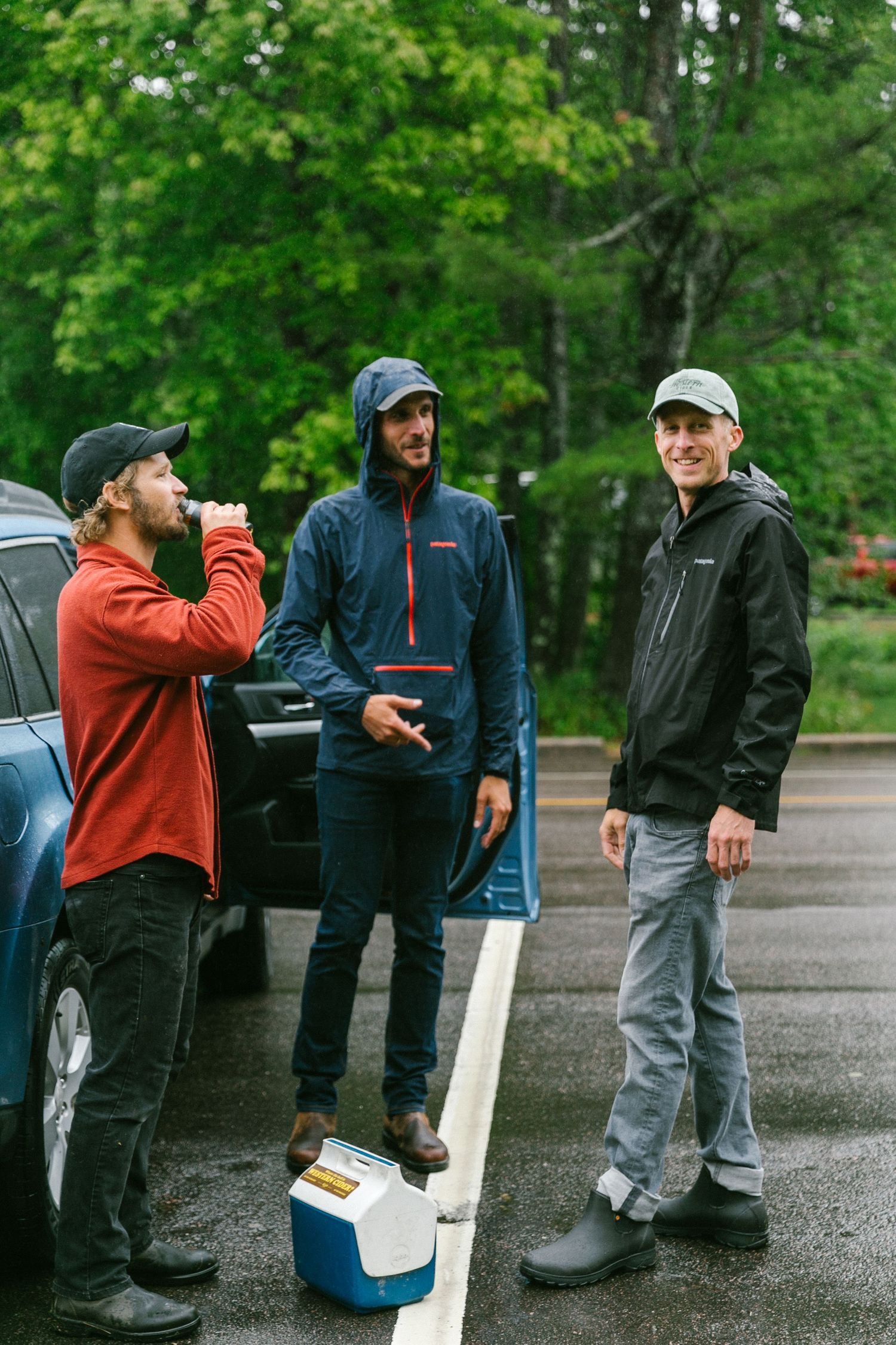 Three friends gather around a cooler in rain gear on a wet day outdoors.