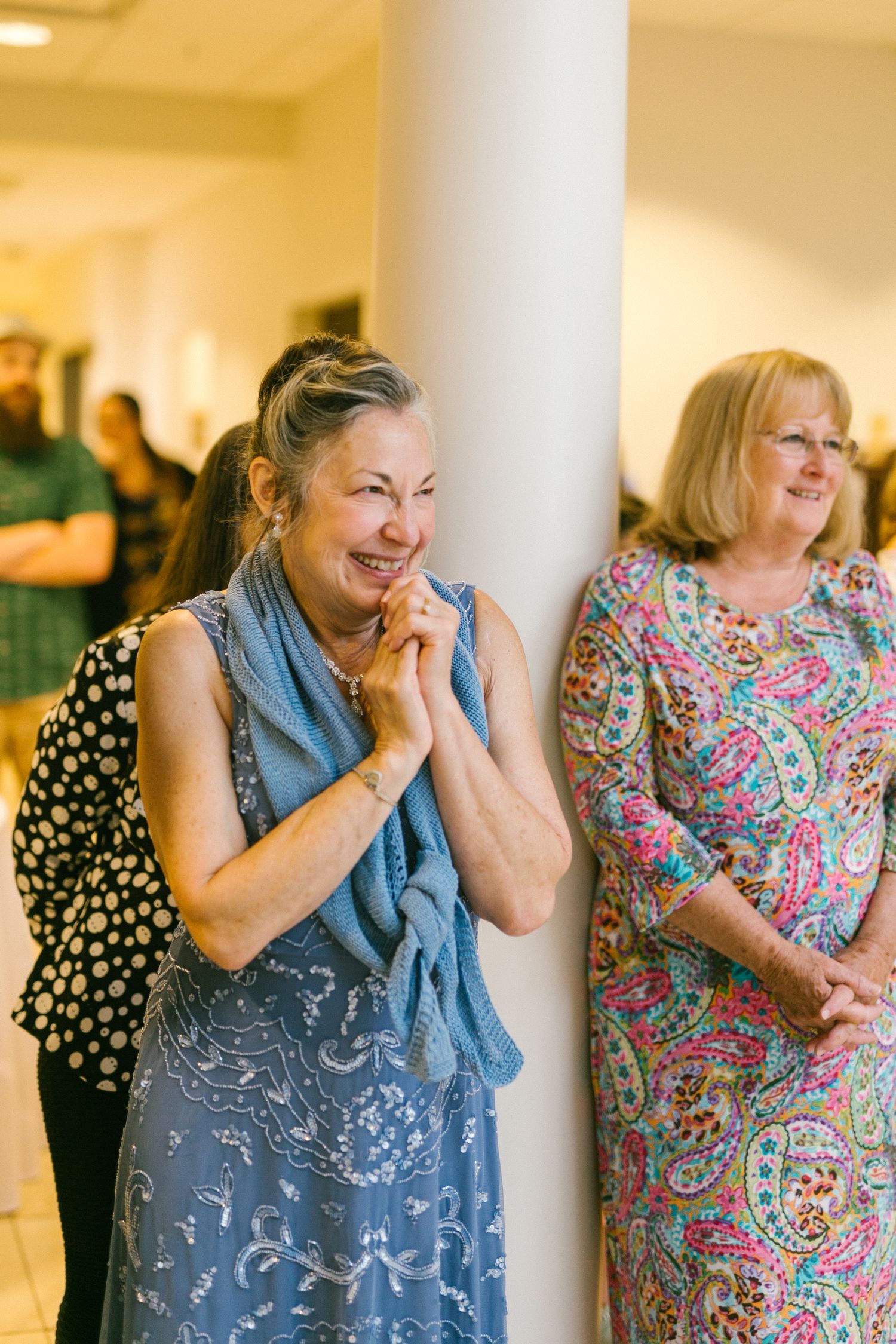 Two women smile joyfully at an indoor event, one wearing a blue patterned dress and another in a colorful paisley print.