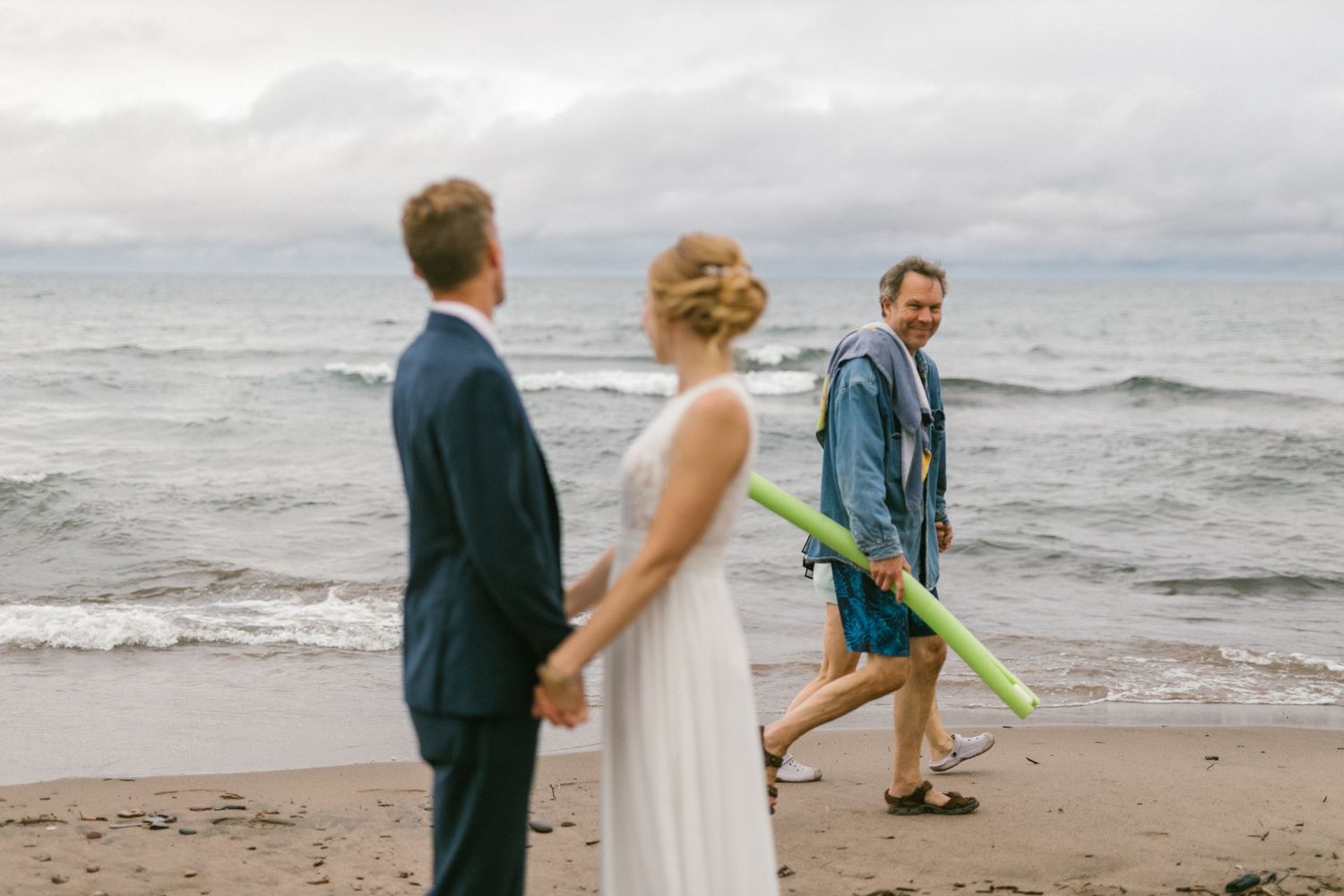 A candid beach wedding moment with a person walking by with a beach toy.