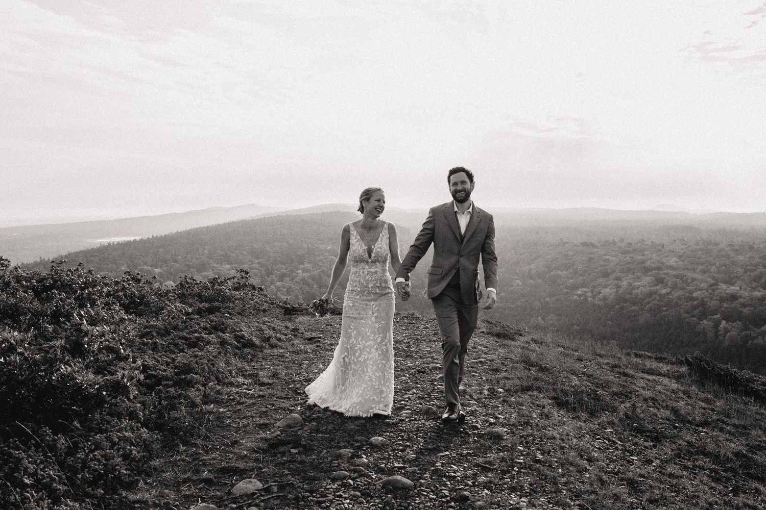 A couple walks hand in hand across a hilltop in black and white, with sweeping mountain views in the distance.