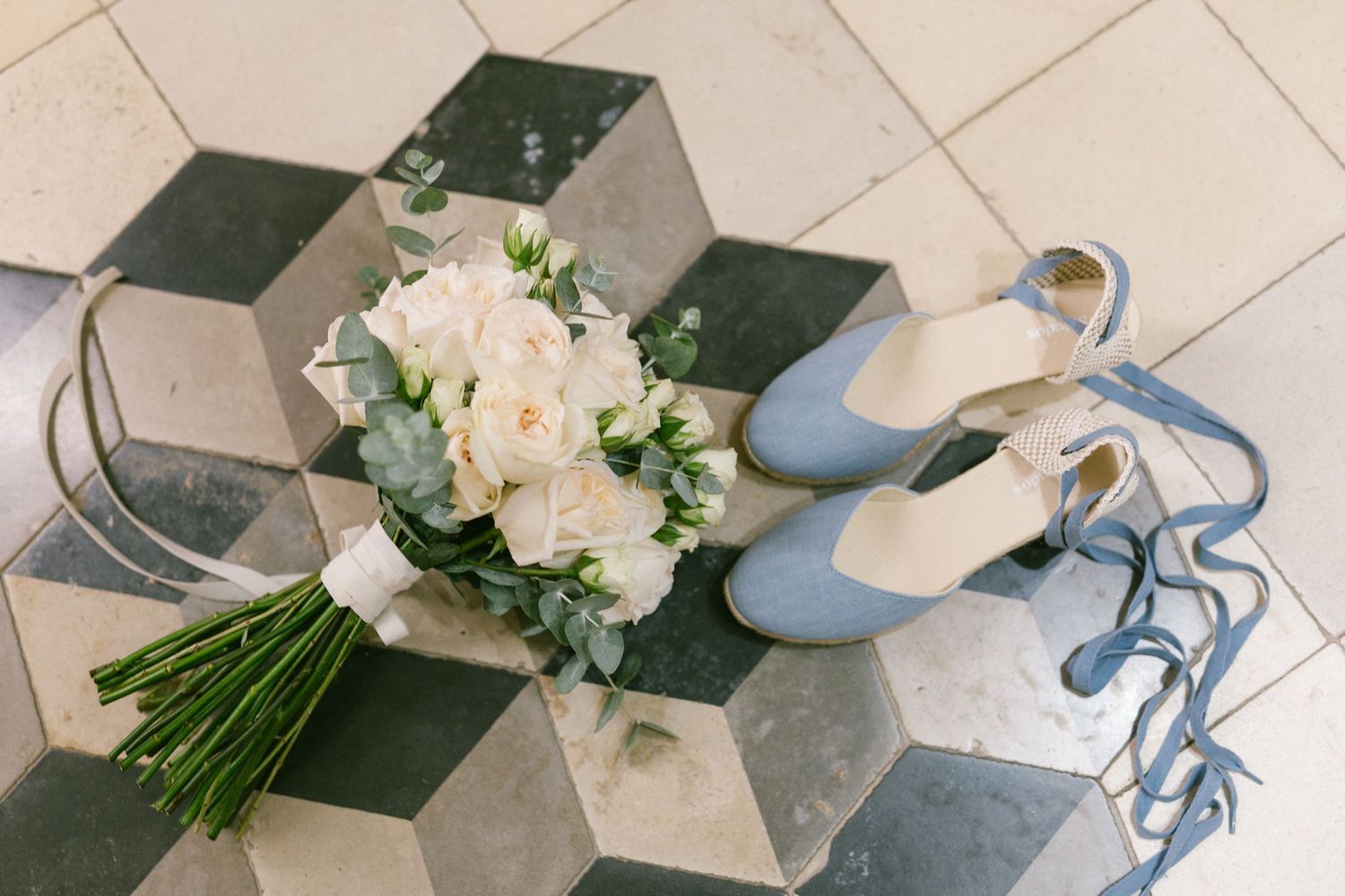 A bridal bouquet of white roses and blue ankle-strap wedding shoes on a vintage hexagon tile floor.