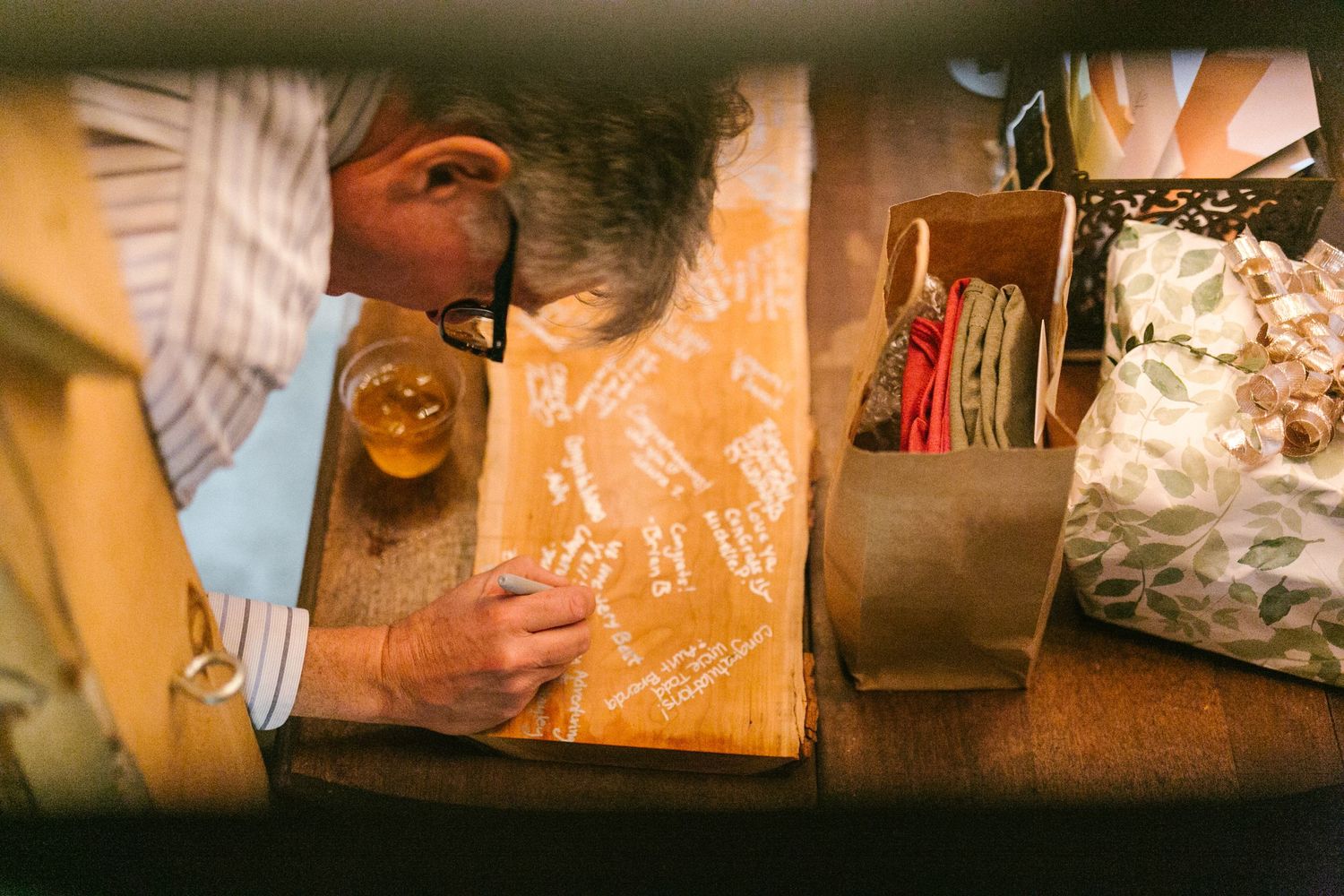 A person enjoys a beer at a rustic wooden bar with signatures etched into its surface.