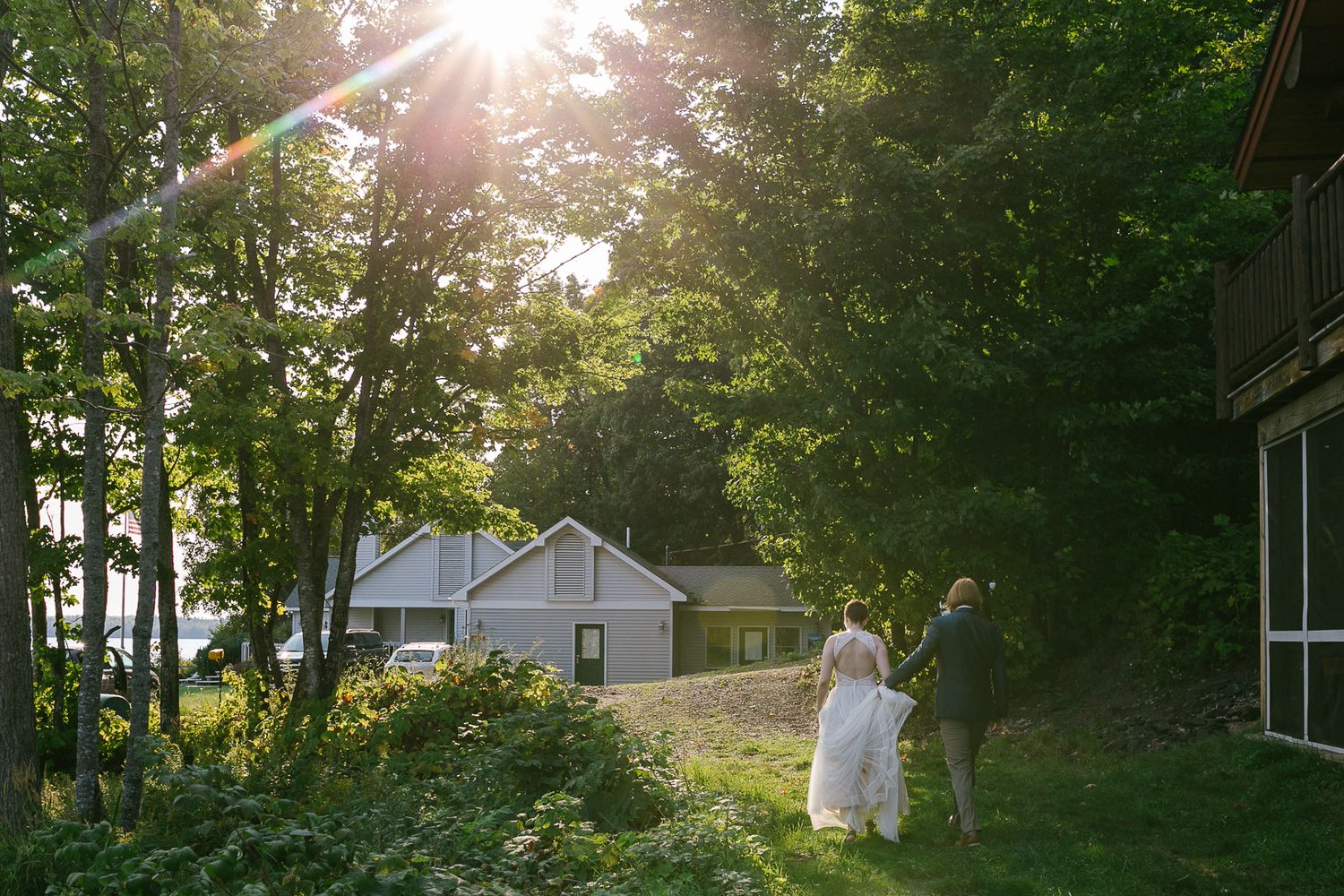 A sunlit path through green trees leads to residential homes at golden hour with lens flare in the background.