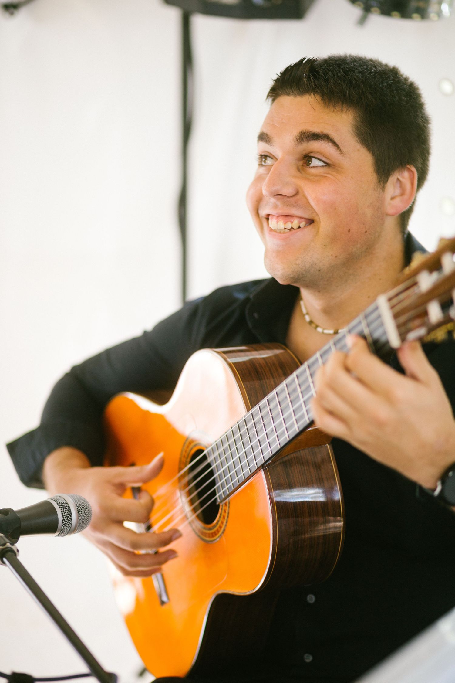 Smiling musician playing a Spanish acoustic guitar with a microphone stand visible in front.