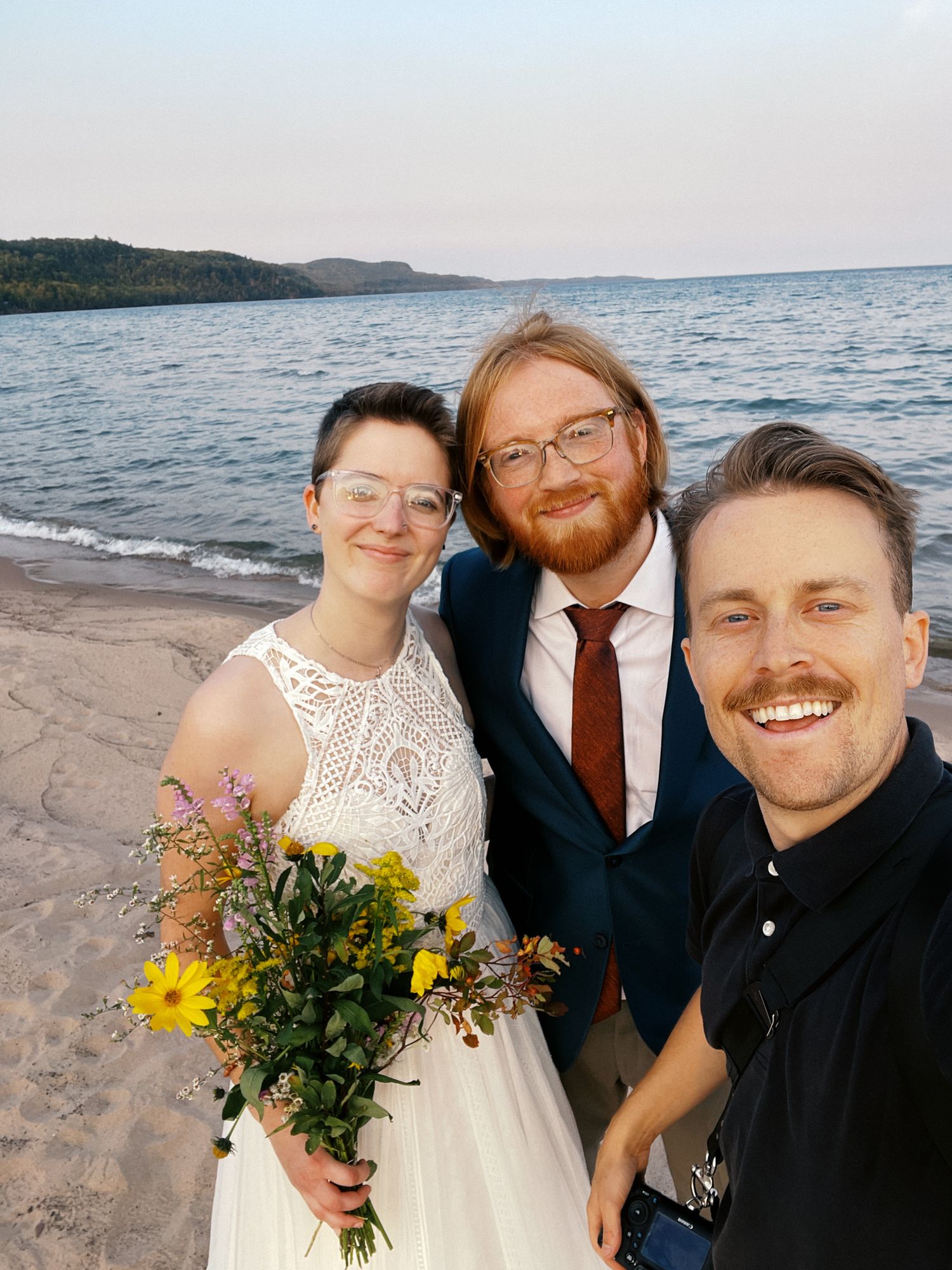 Couple in wedding attire with bouquet poses for beach selfie with their photographer at sunset by a lake.