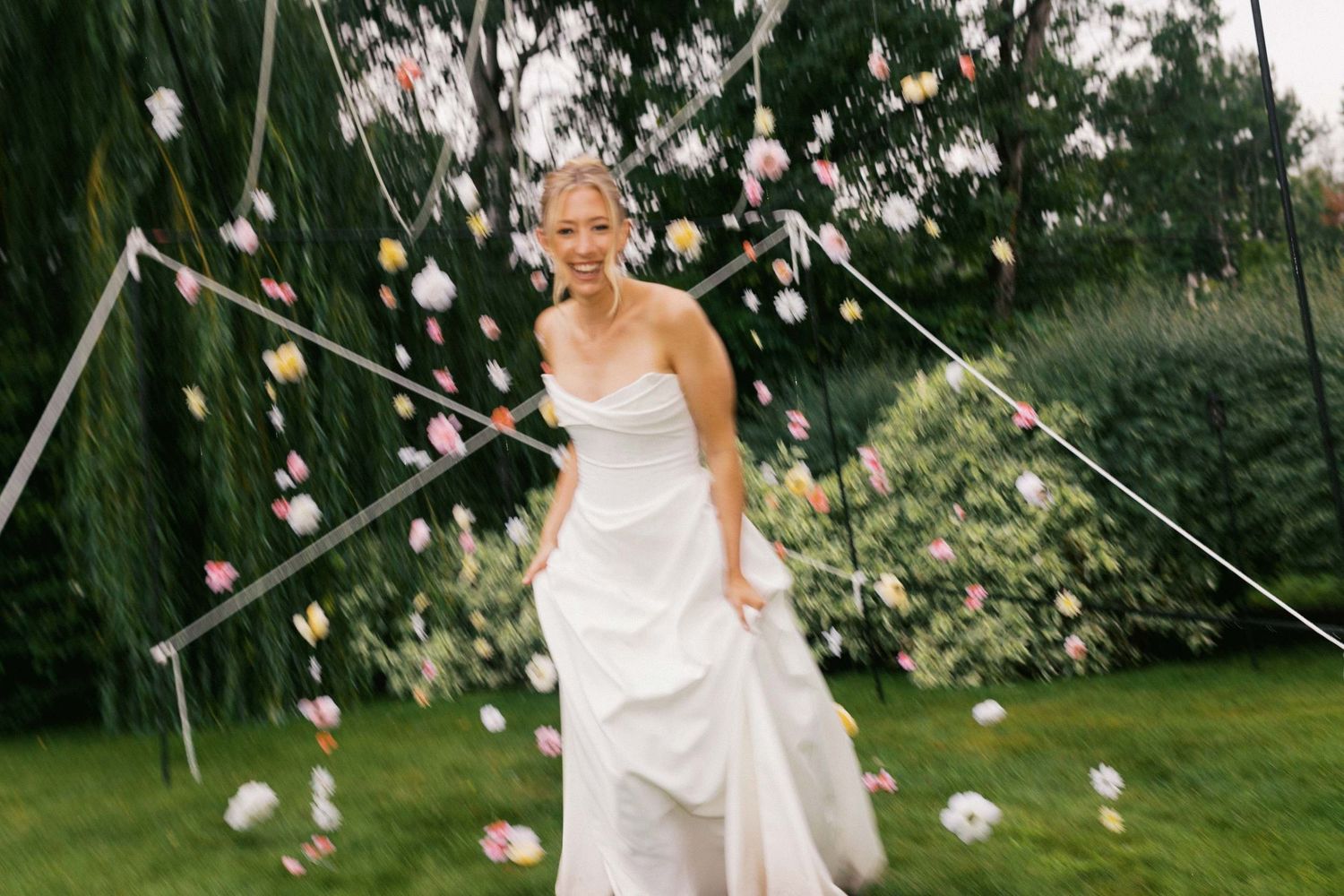 A bride in a white wedding dress twirls playfully in a garden setting surrounded by pink roses and greenery.