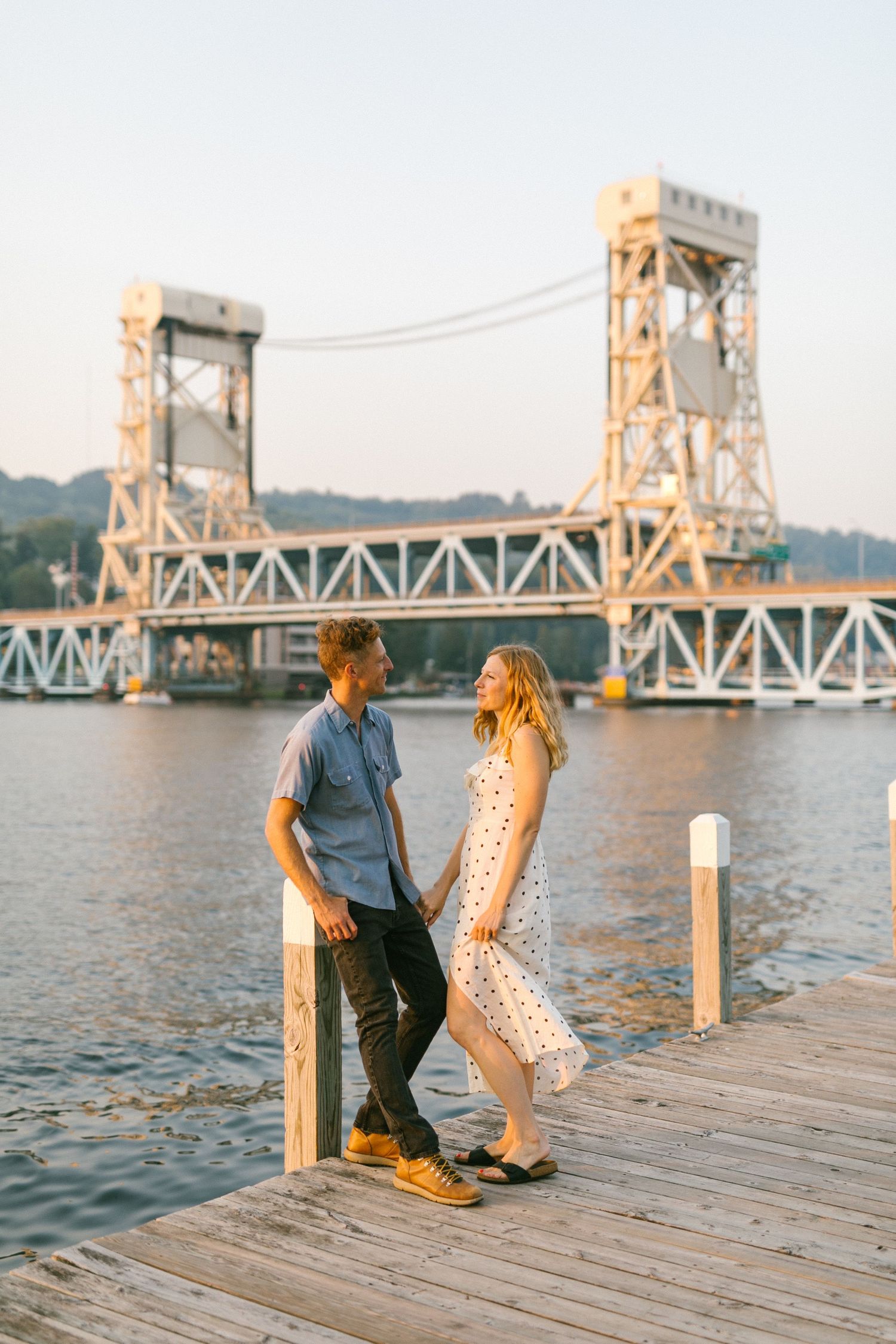 Couple in casual attire playing and dancing on a wooden pier with a bridge in the background.