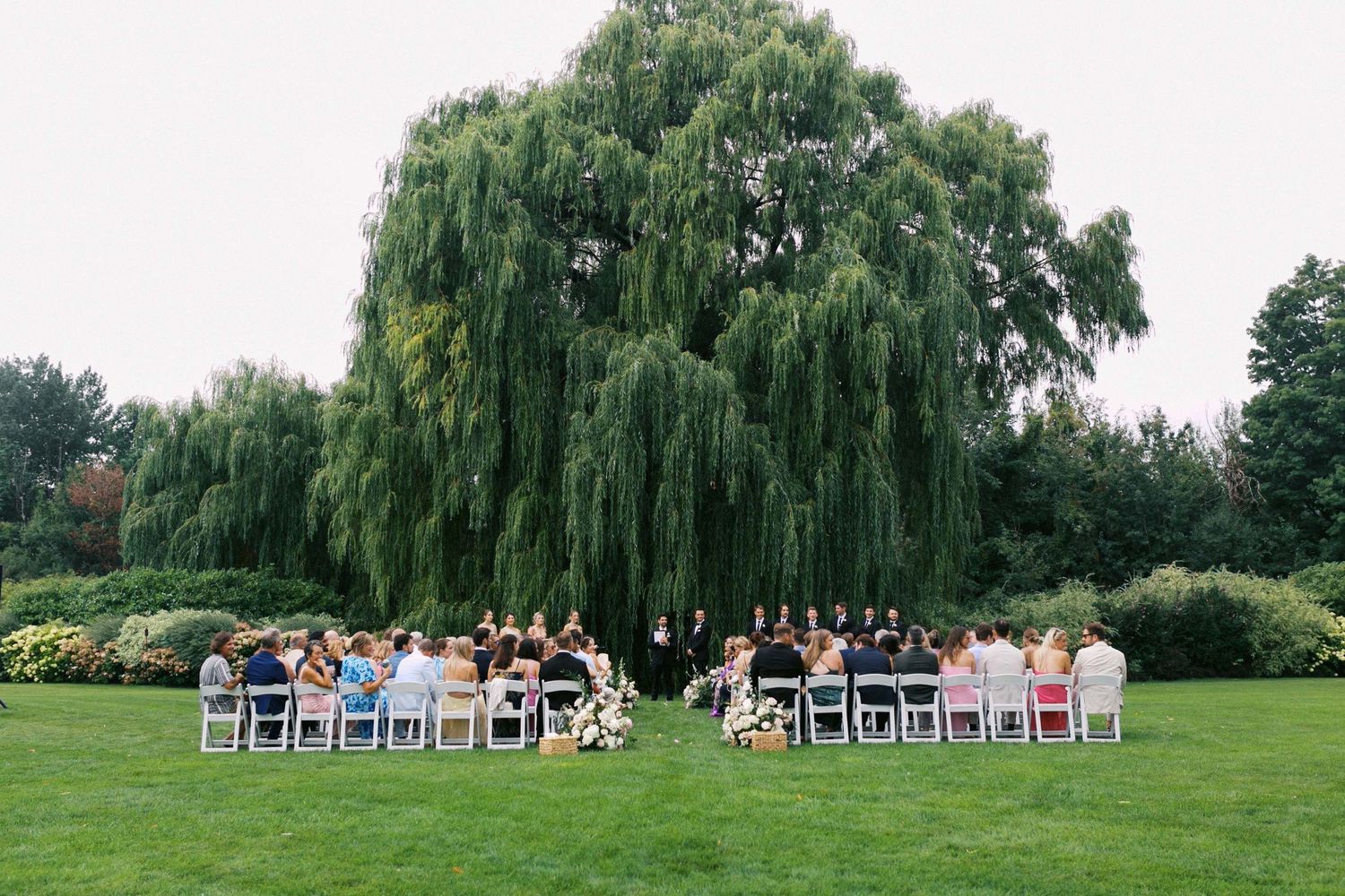 Outdoor wedding ceremony setup with white chairs arranged in rows on a lush green lawn surrounded by willow trees.