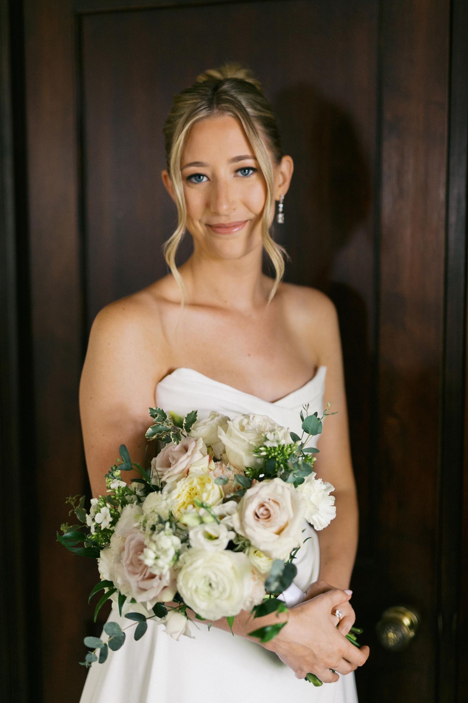 A bride in a white strapless gown holds an elegant bouquet of blush roses, white flowers and eucalyptus greenery.