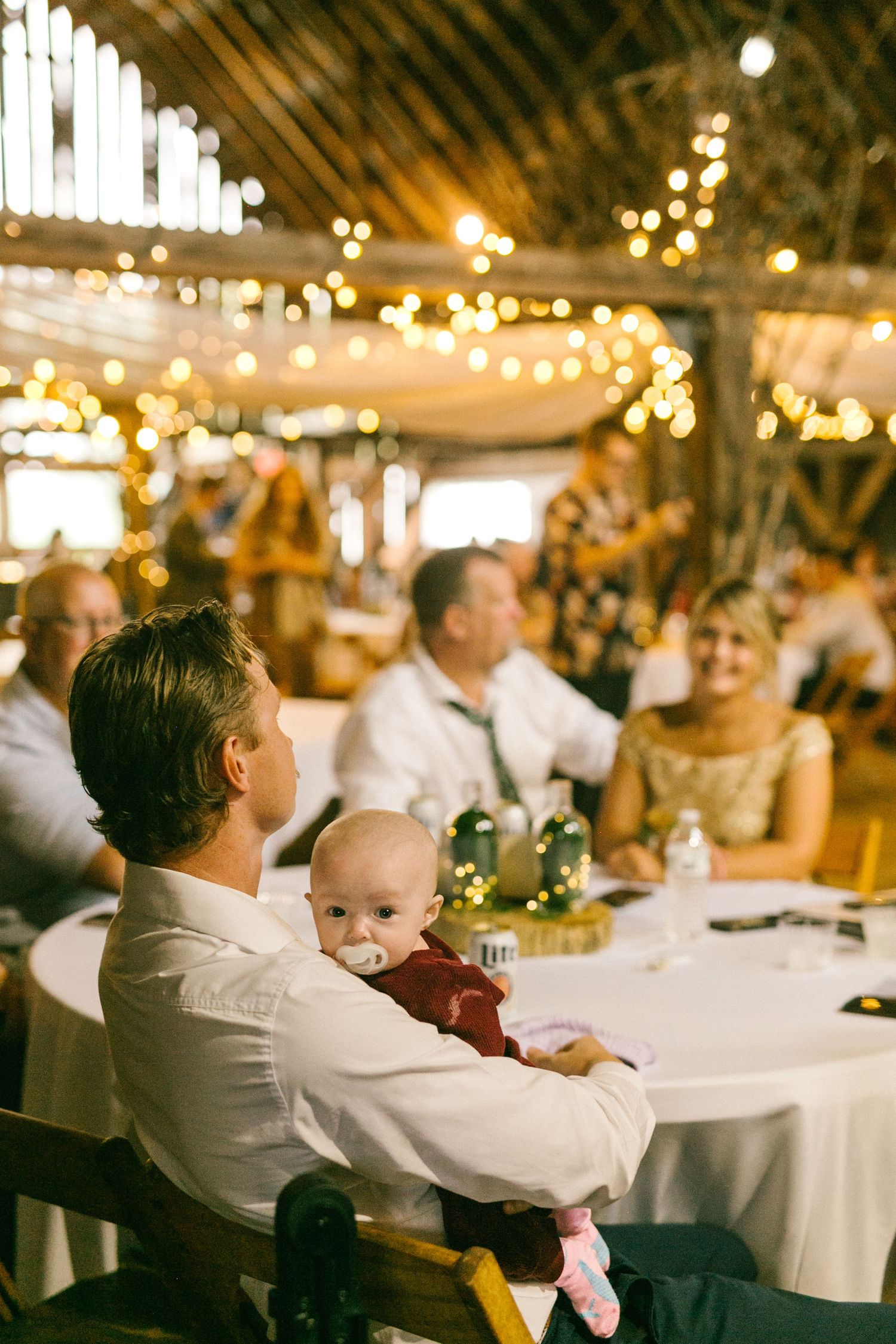 A baby sits with a guest at an evening wedding reception decorated with warm glowing string lights.