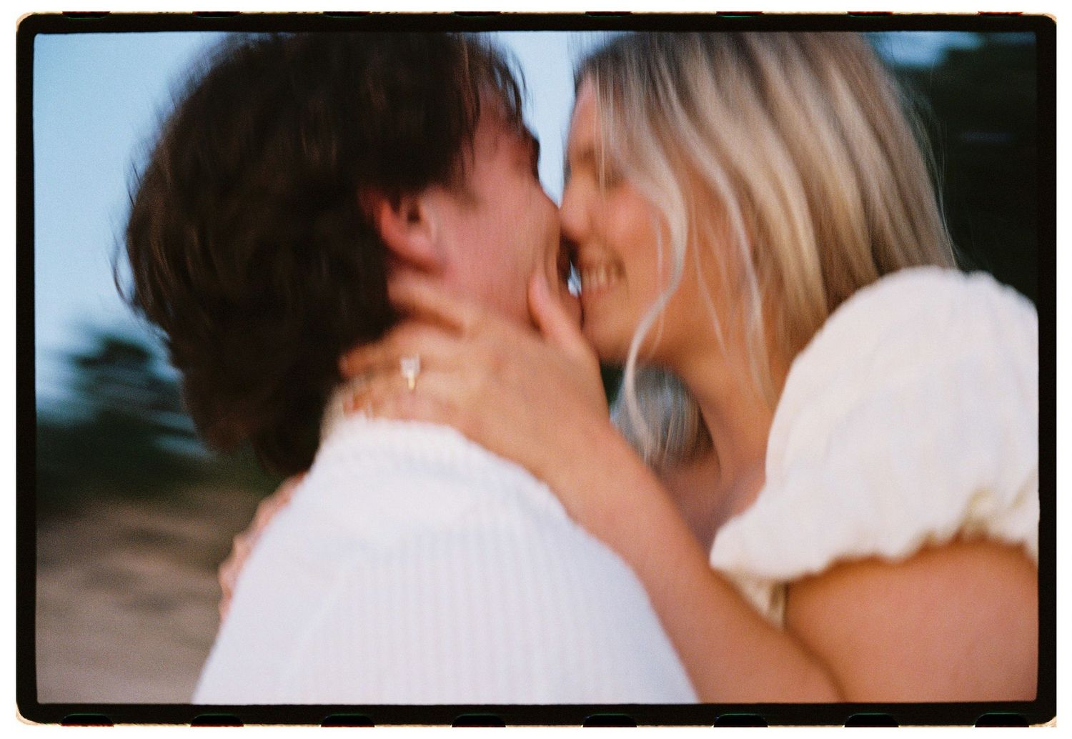 Romantic couple sharing a tender kiss outdoors, woman with blonde hair wearing white dress and ring.
