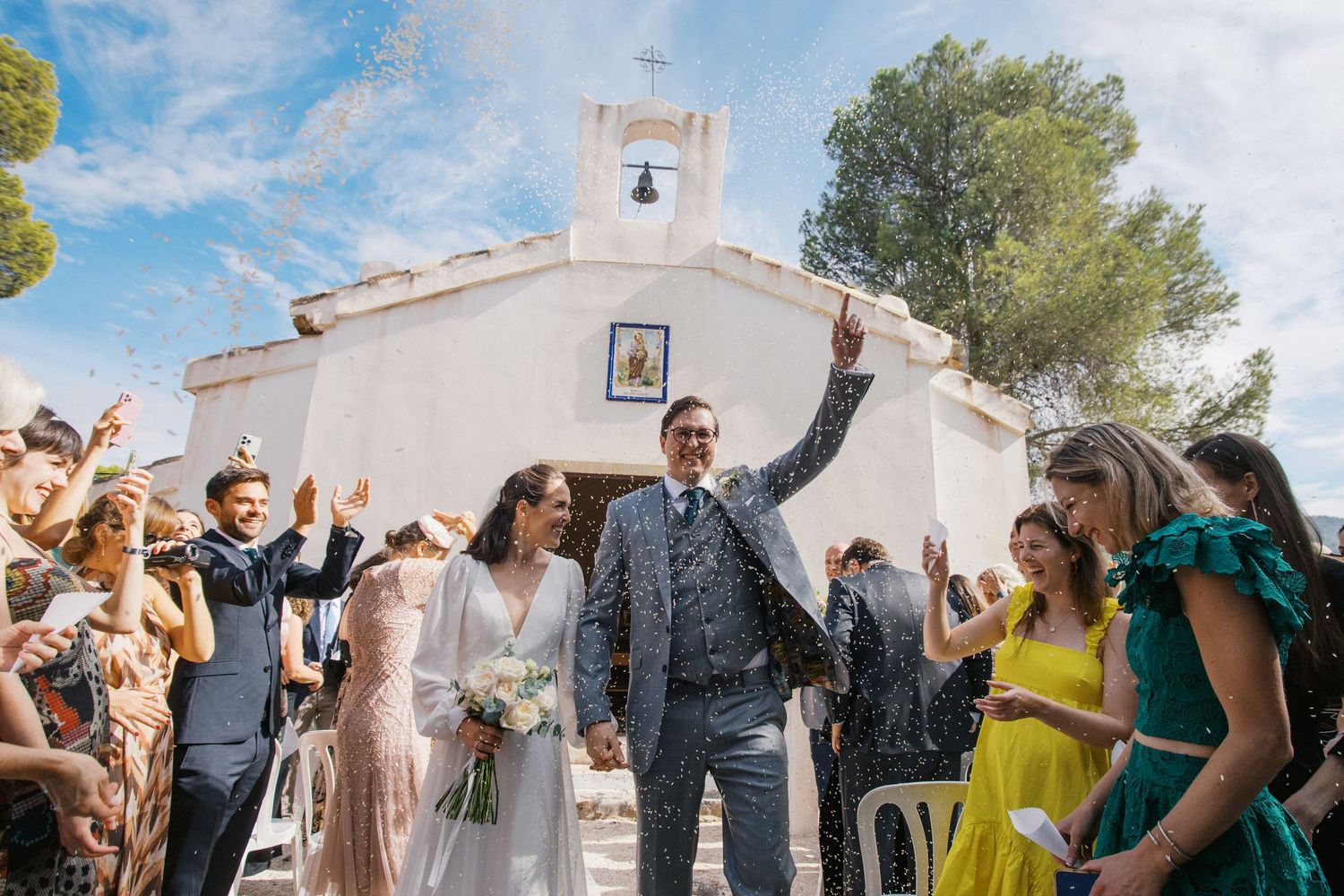 Bride and groom exit a charming white chapel surrounded by cheering wedding guests on a sunny day.