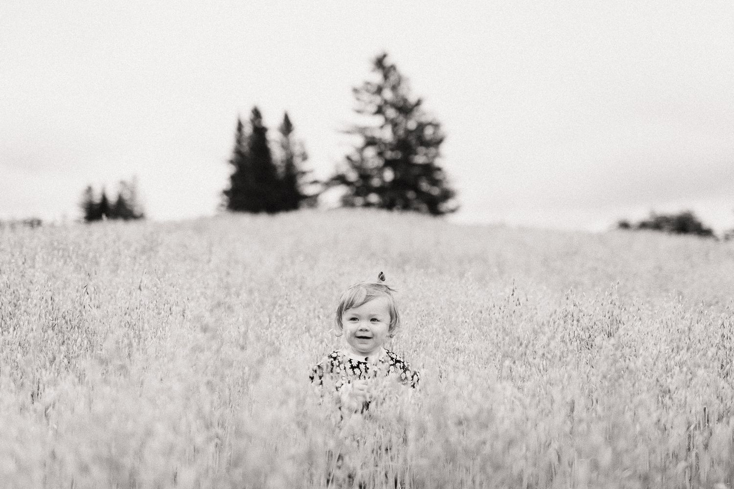 Black and white photo of a happy toddler standing alone in a tall grass field with trees behind.