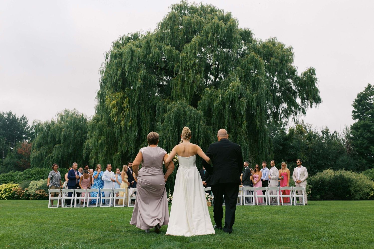 Wedding party stands in a row on lush green lawn with willow trees in the background during outdoor ceremony.