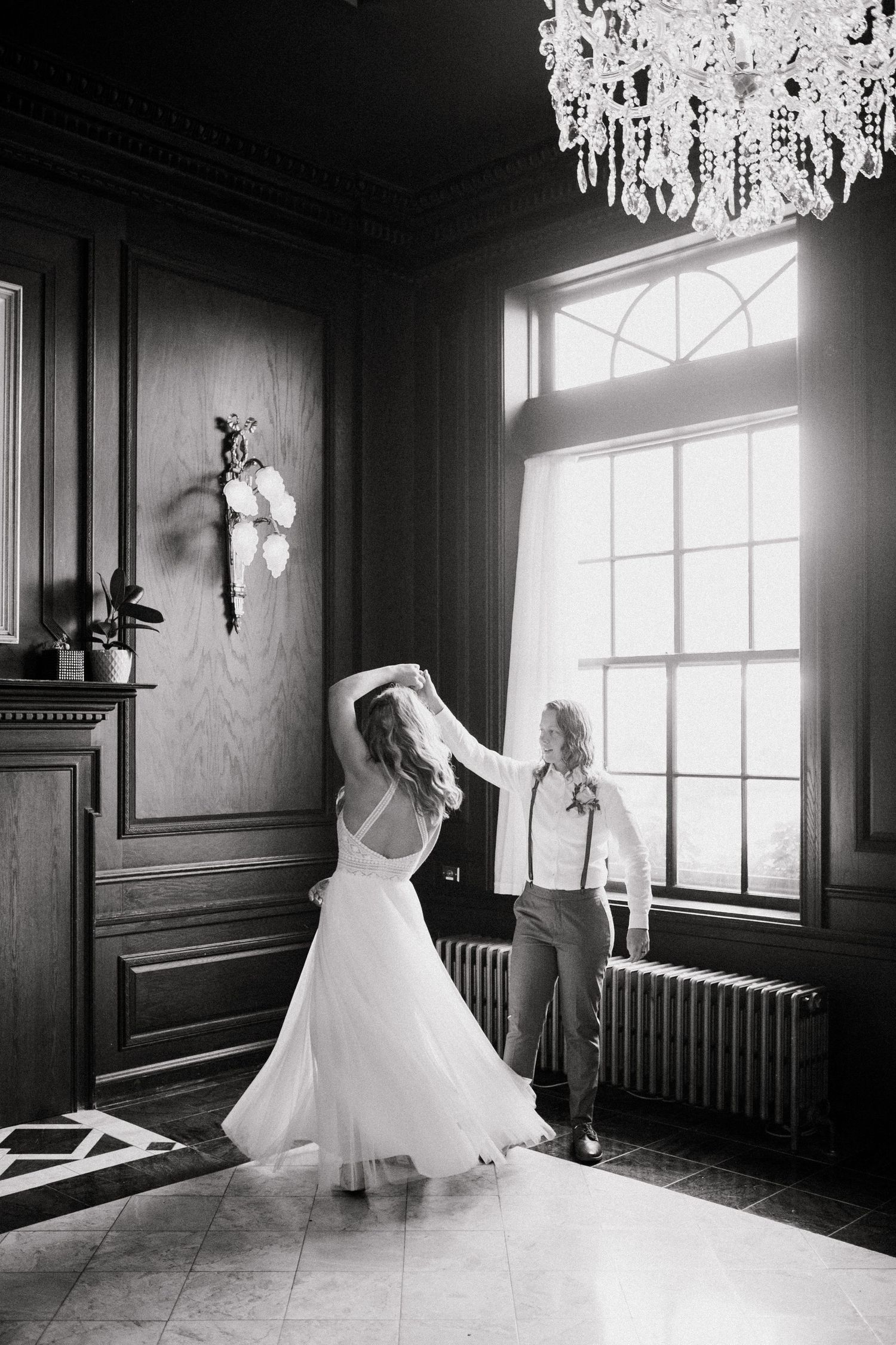 A black and white photo of a wedding dance scene in an elegant room with a crystal chandelier and large windows.
