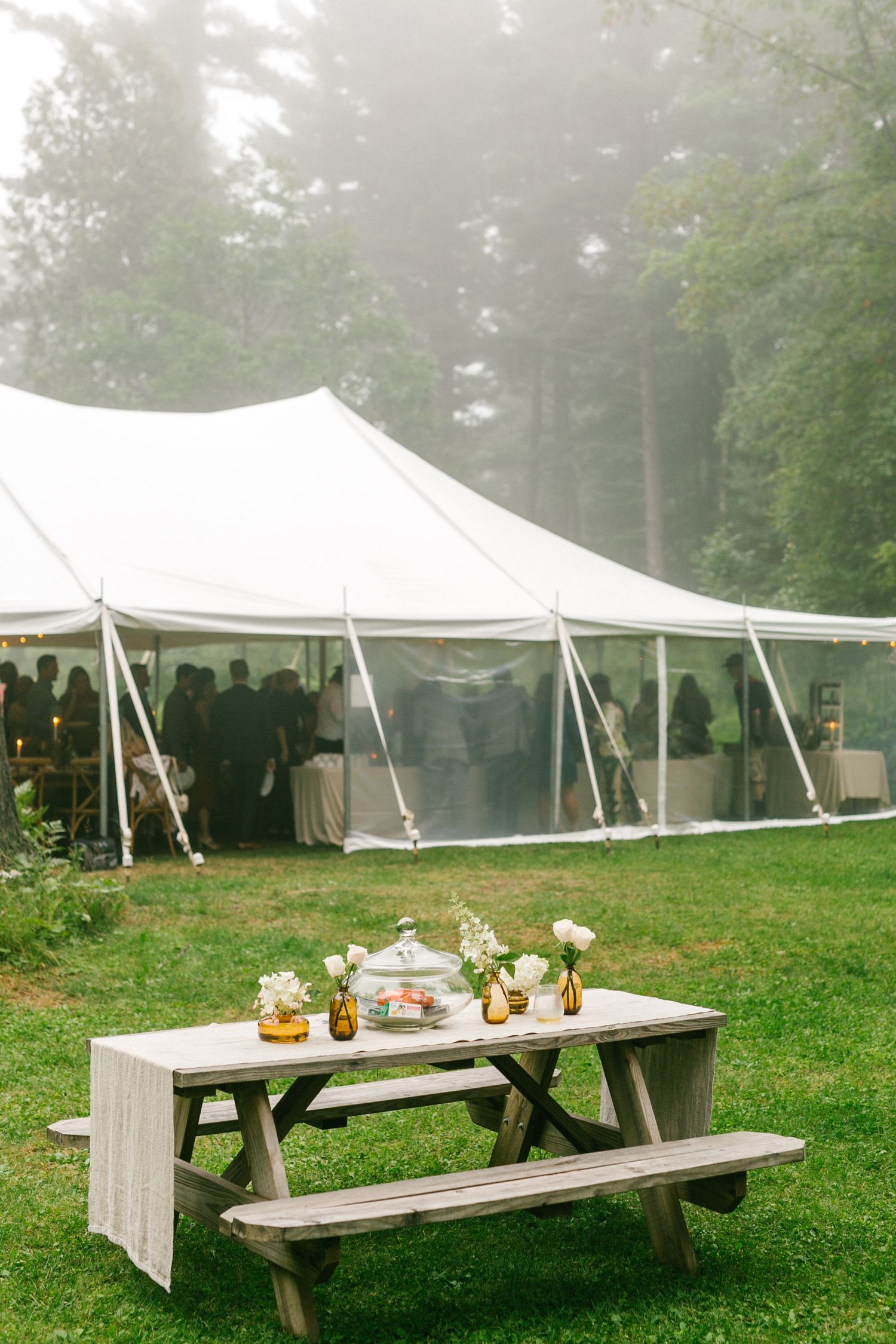 White wedding tent setup in a misty forest clearing with rustic picnic tables decorated with floral centerpieces.