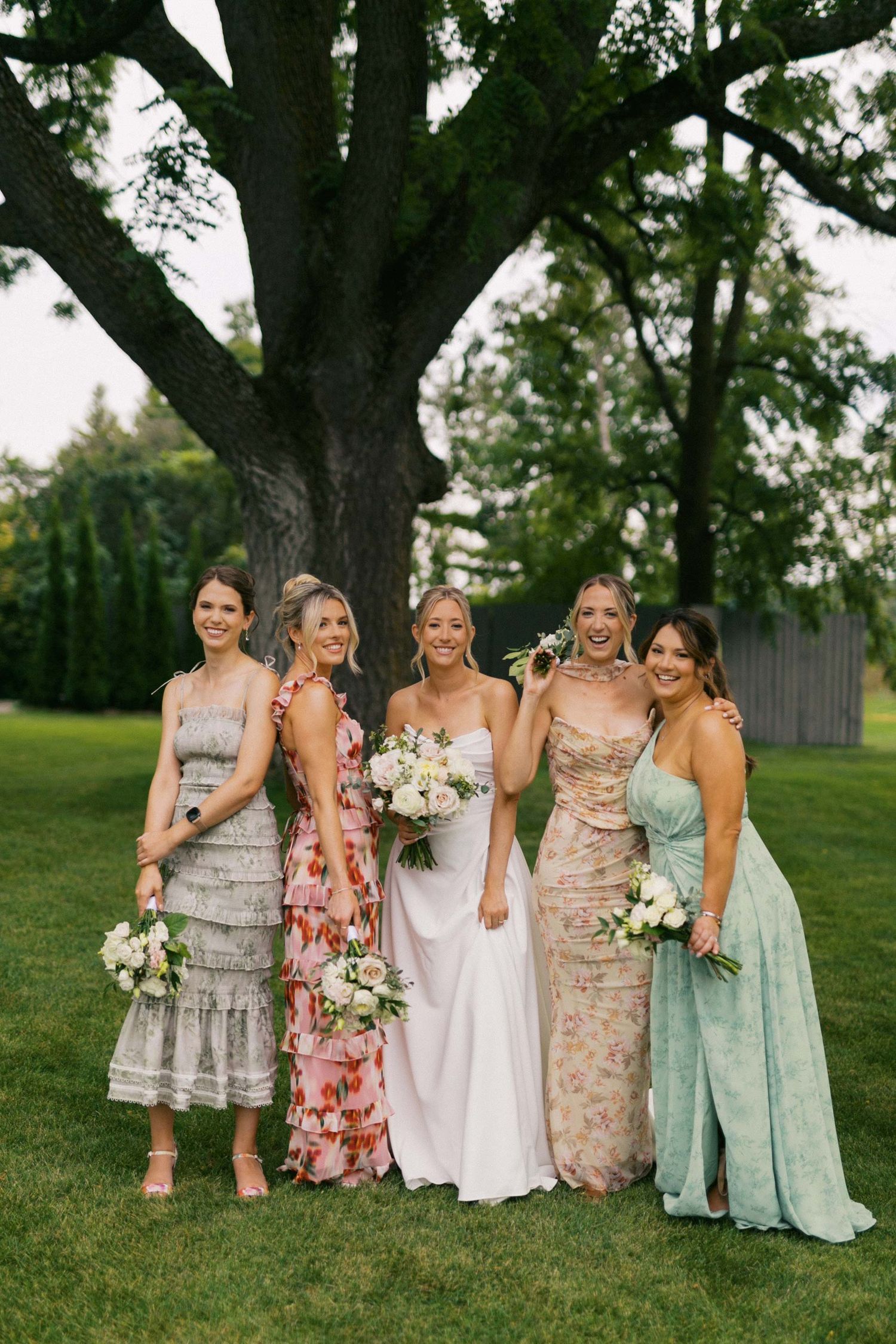 Bridesmaids in pastel floral dresses and soft mint gowns pose together on a grassy lawn at an outdoor wedding.
