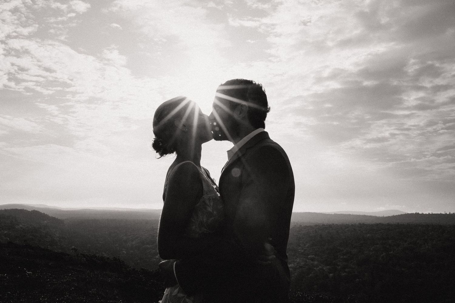 Two silhouetted figures share an intimate moment against a dramatic sunset sky over rolling hills in black and white.