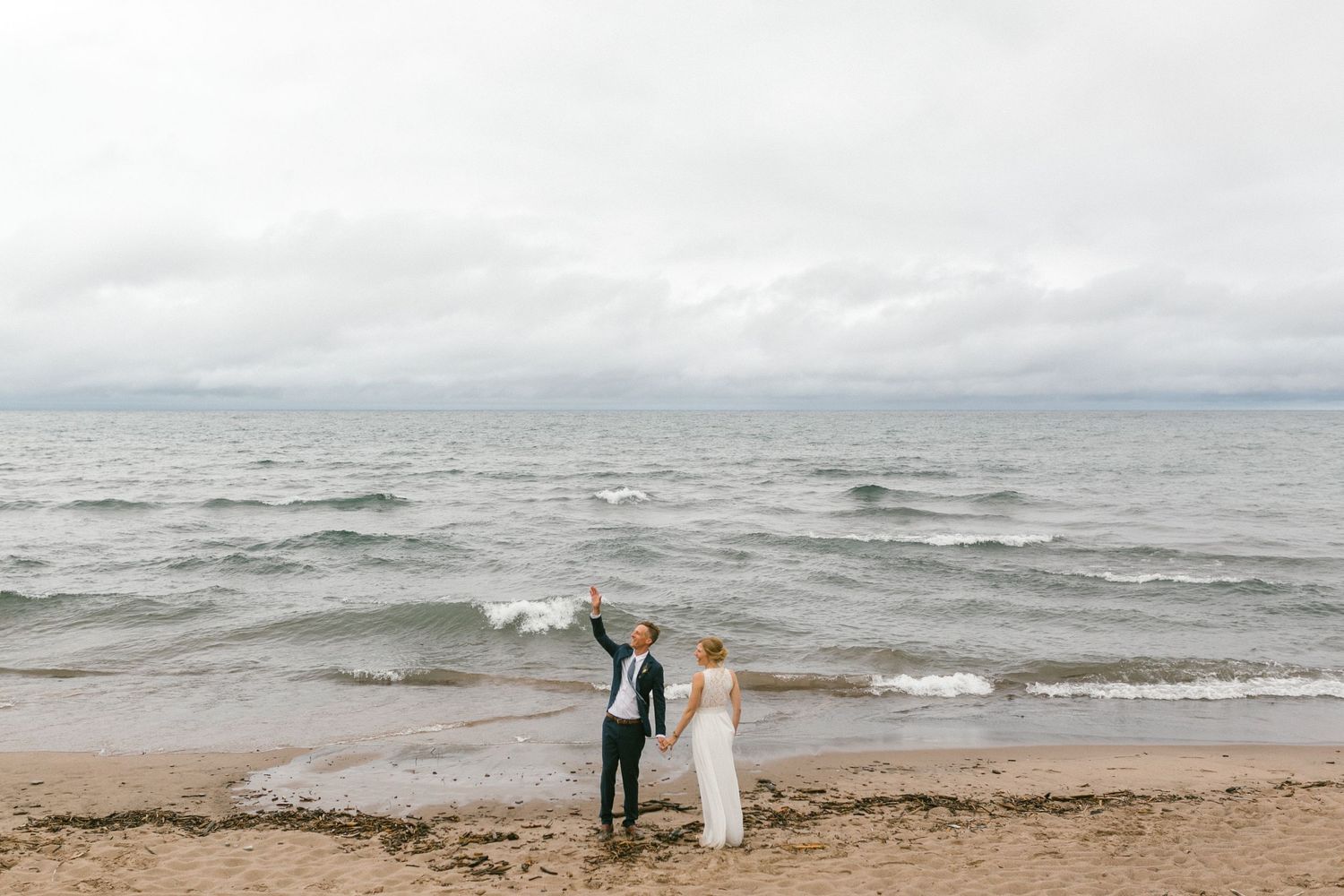 Two figures stand at the edge of a stormy beach with waves crashing behind them on an overcast day.