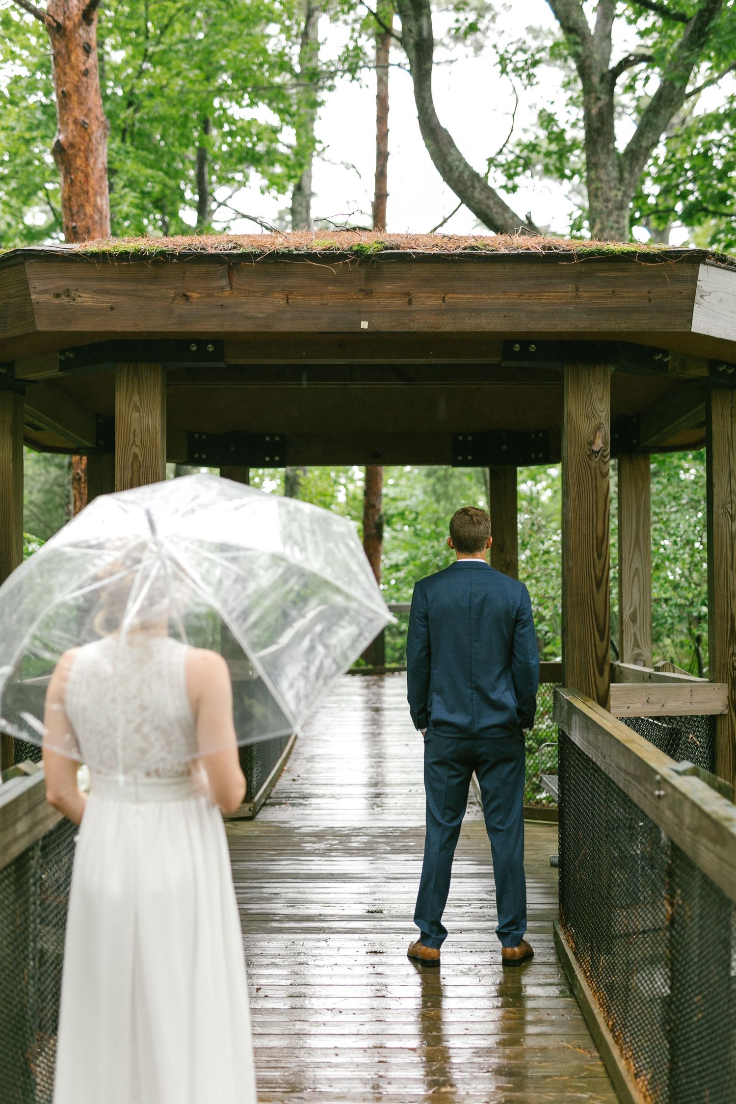 A couple stands on a rainy wooden walkway with a clear umbrella in a forested setting.