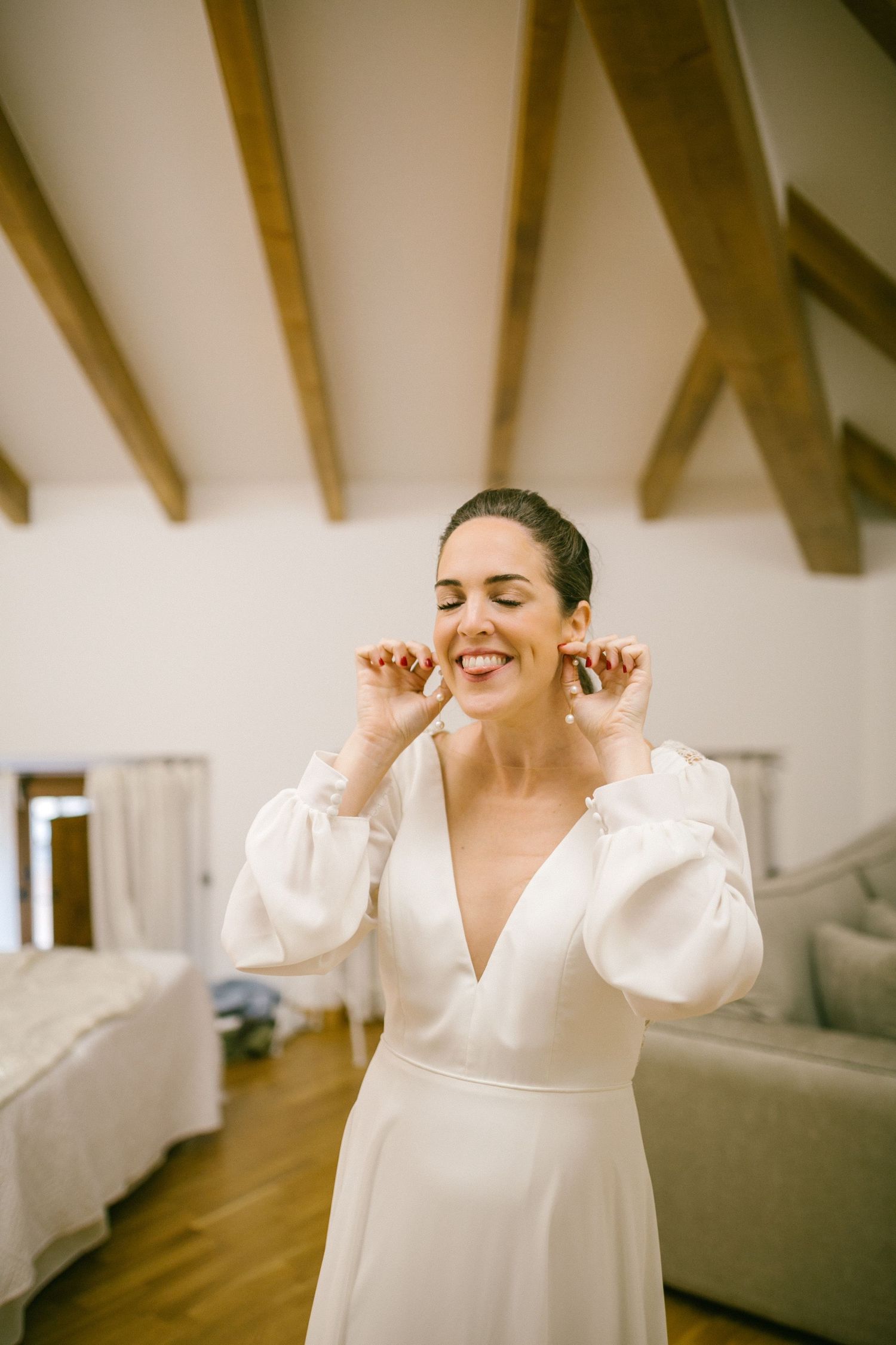 Bride in elegant white long-sleeve wedding dress smiles joyfully with closed eyes in a room with exposed wooden beams.