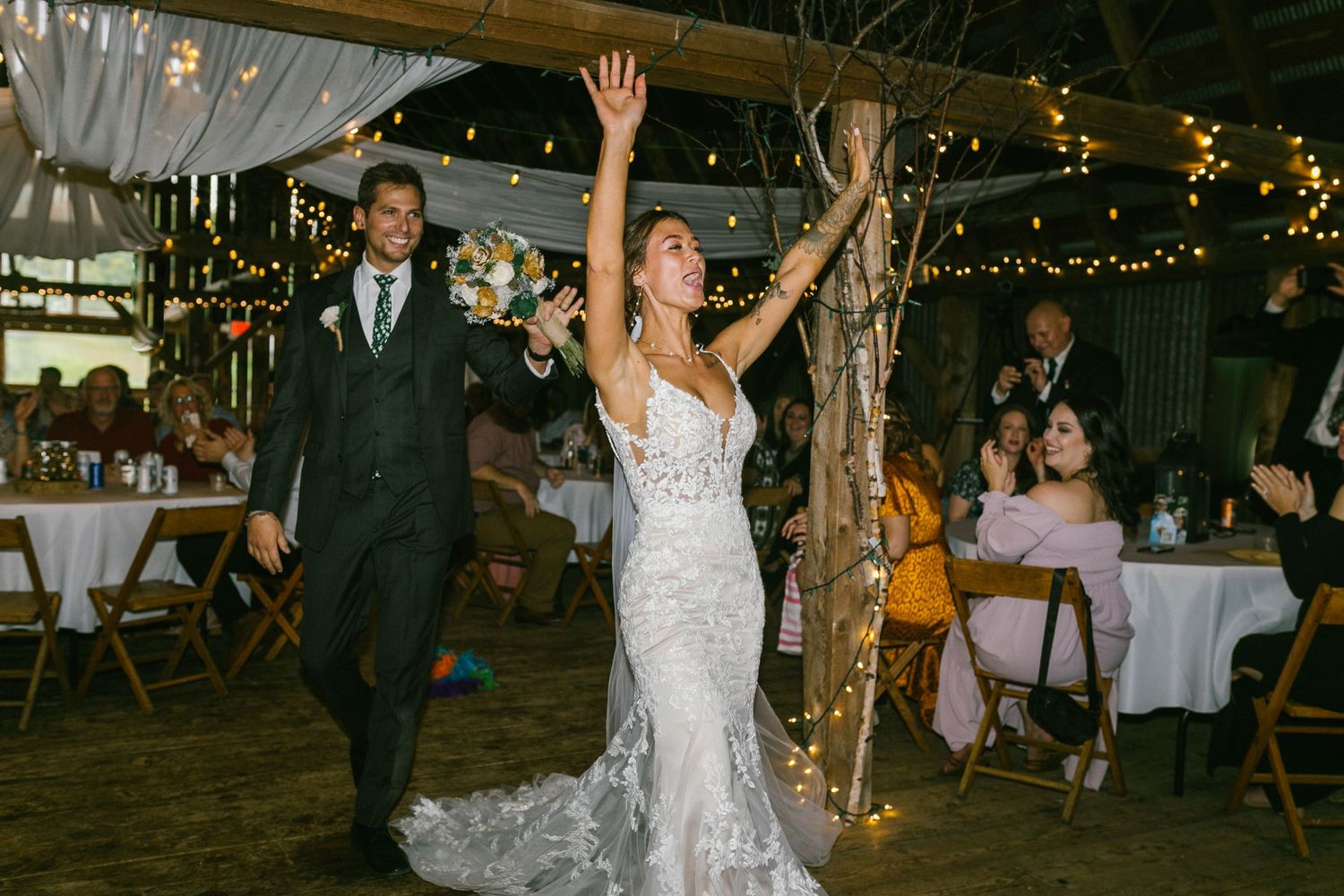 Newlyweds make their grand entrance at rustic barn reception with string lights and white decor.
