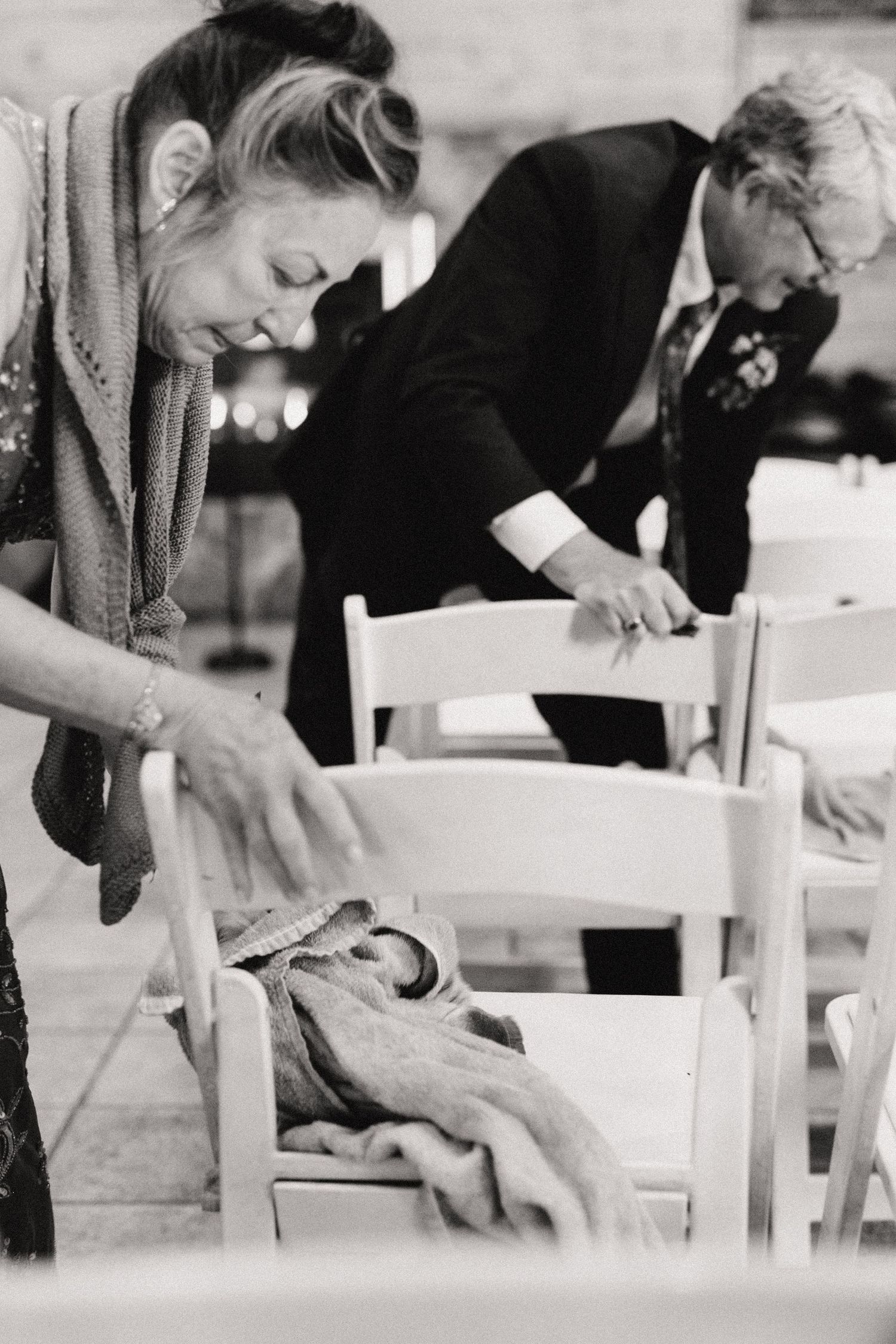 Black and white vintage photograph showing two people arranging white wooden chairs in an interior space.