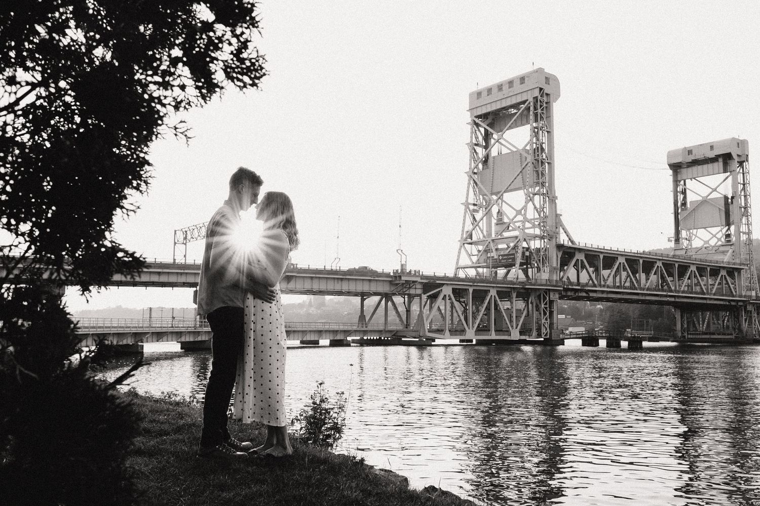 Silhouetted couple against sunburst by historic bridge at sunset in black and white.
