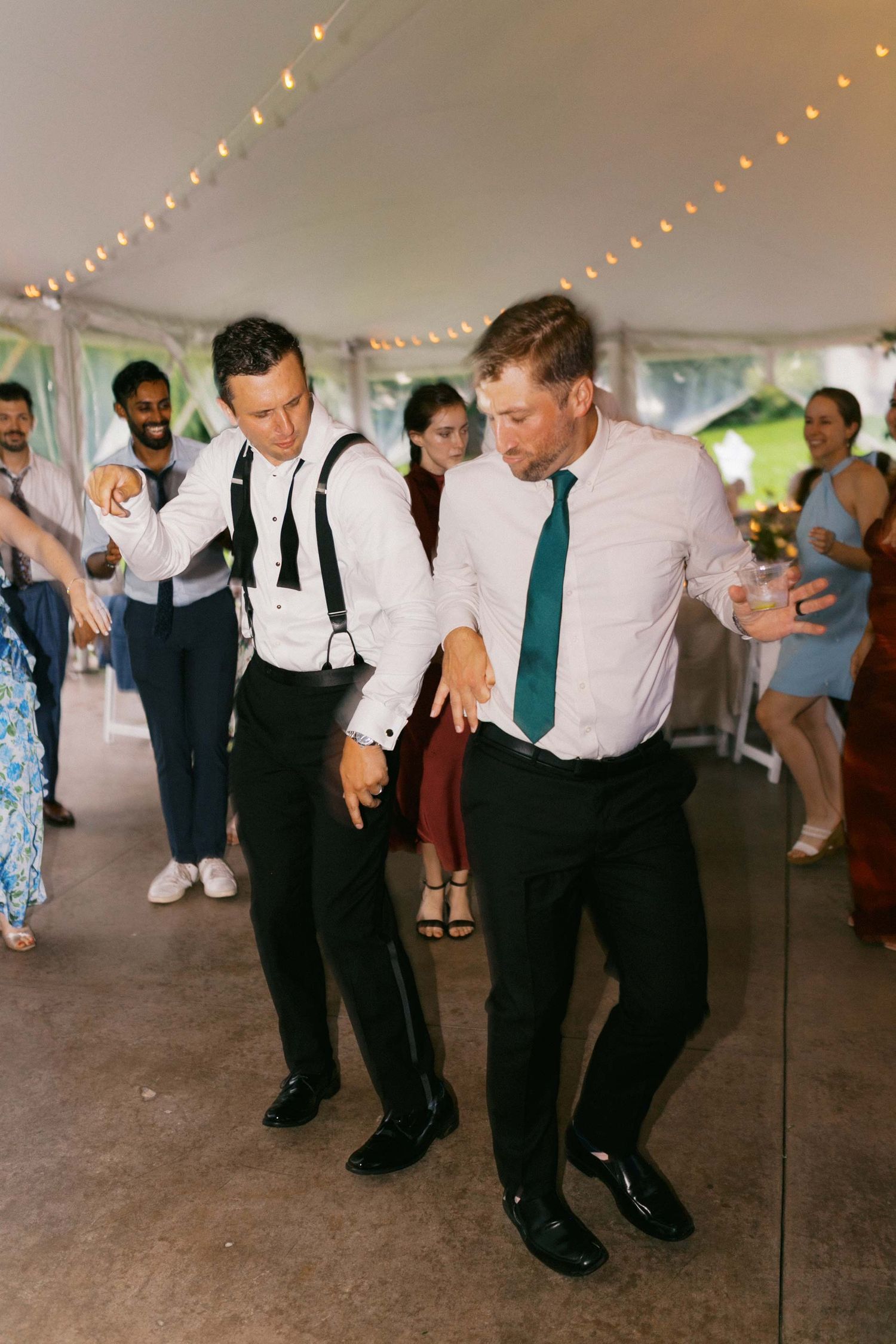 Wedding guests dancing in a line on a covered outdoor dance floor with string lights overhead.