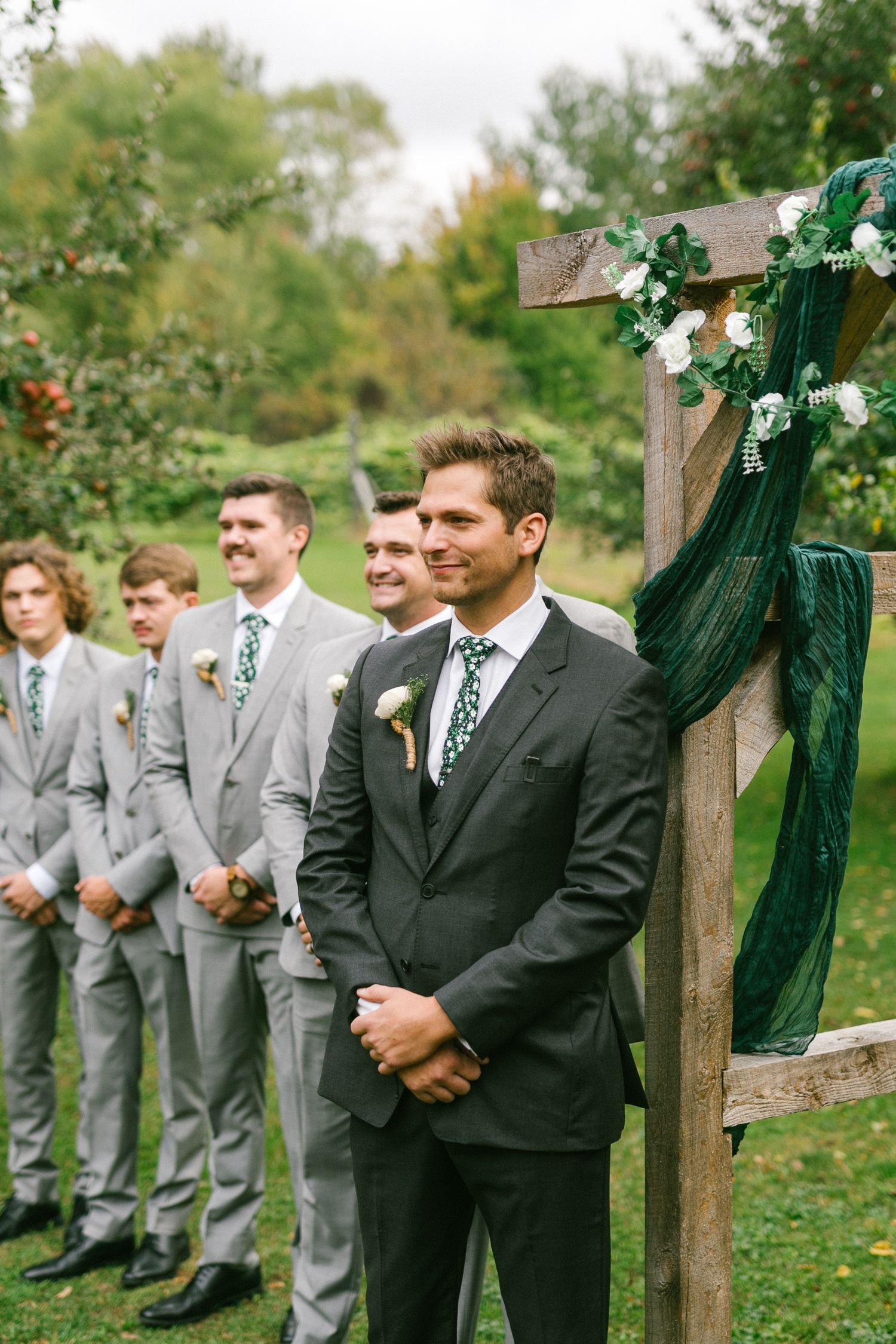 Groomsmen in grey and black suits stand at attention during an outdoor wedding ceremony.