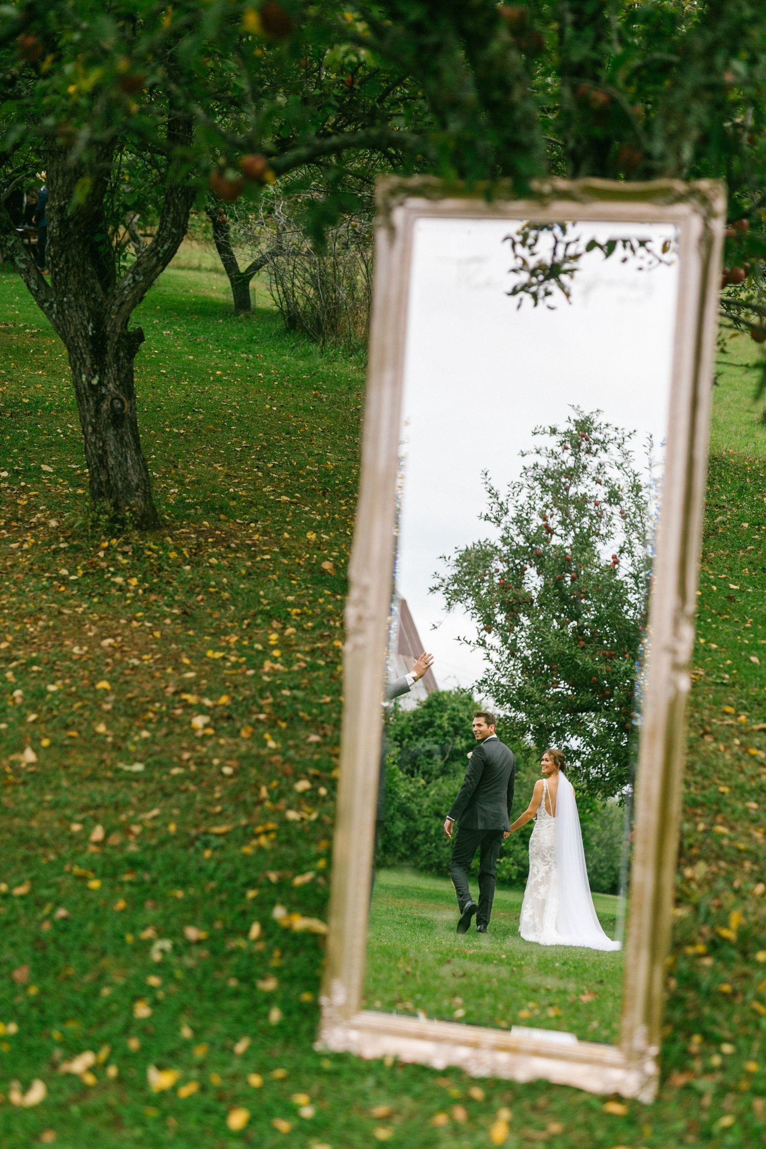A tall ornate mirror stands in a garden reflecting a bride and groom and autumn leaves scattered on the green lawn.
