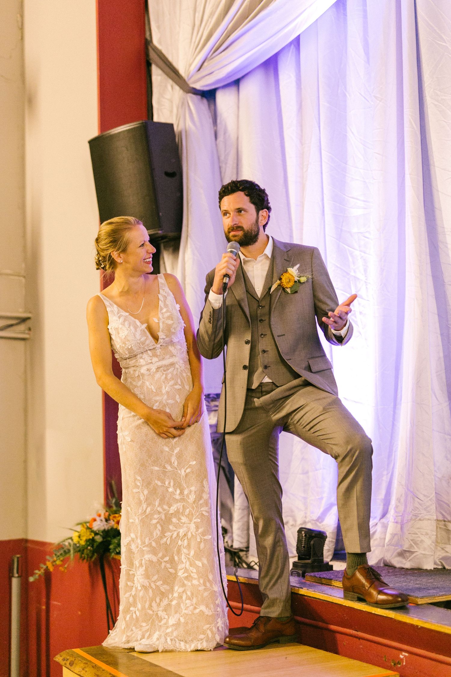 Newlyweds share a moment on stage as the groom speaks into a microphone during their wedding reception.