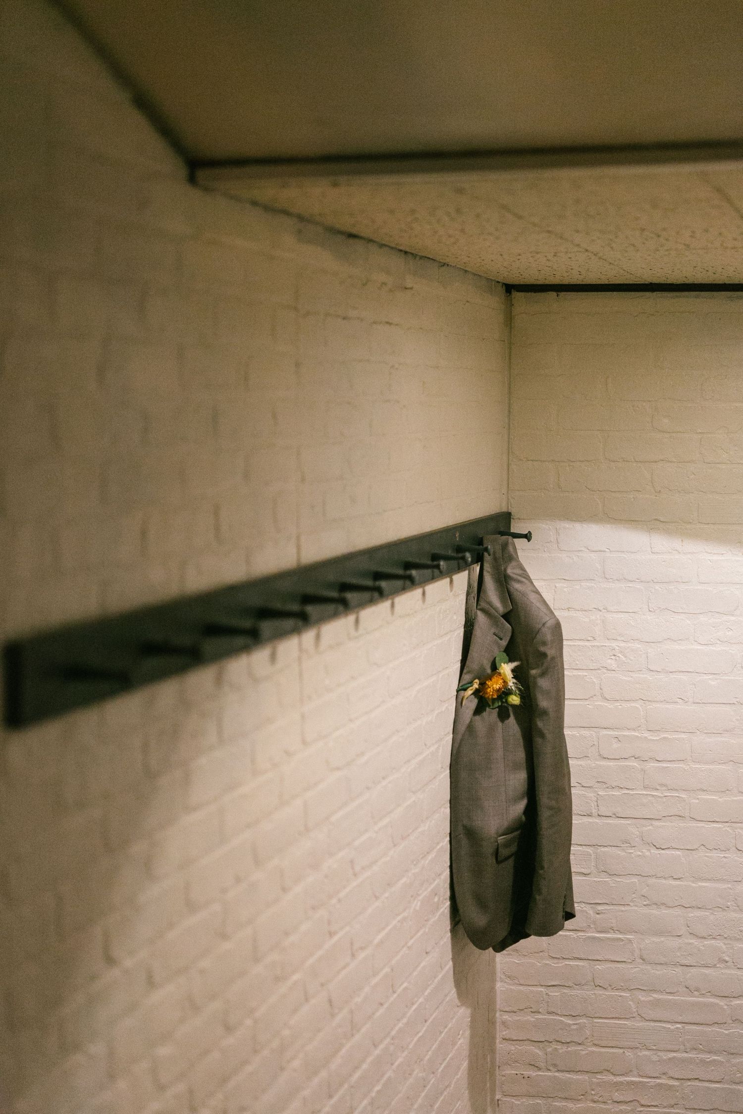 A gray suit jacket with a yellow boutonniere hangs on a coat rack against a white brick wall.