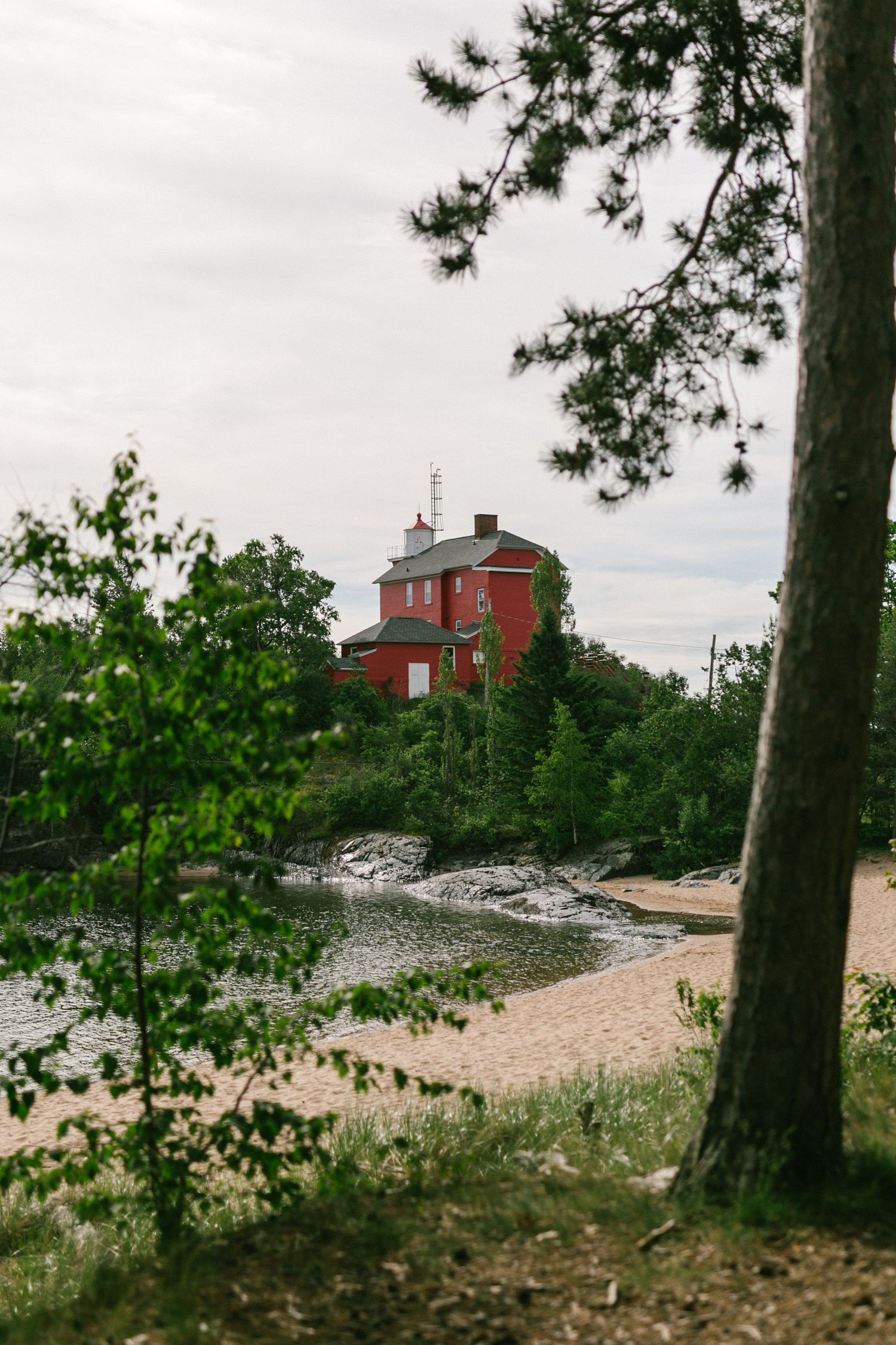 A red lighthouse overlooking Lake Superior stands among trees on a rocky shoreline with a sandy beach below.